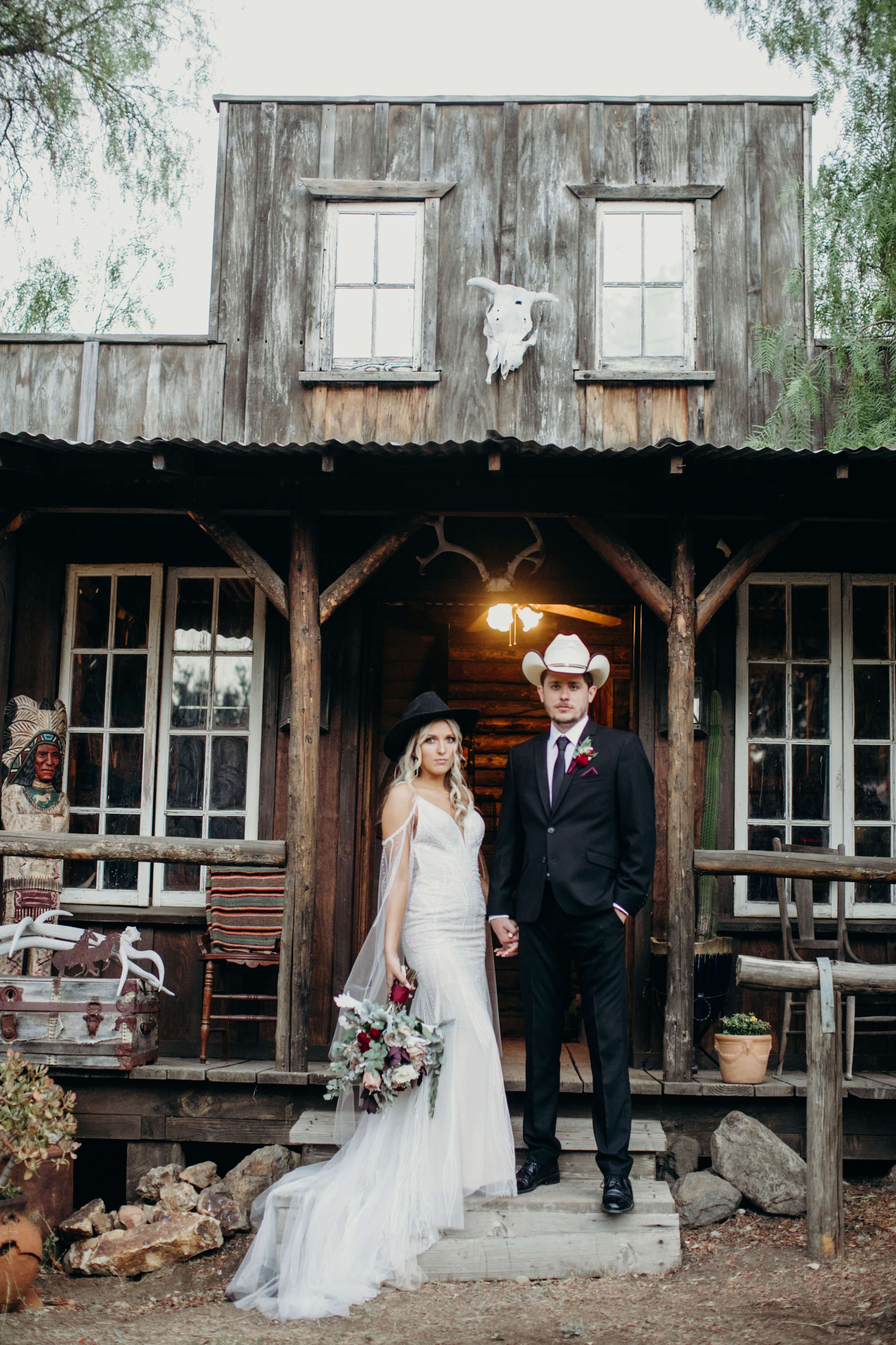 A bride and groom stand together in front of a rustic wooden cabin, holding hands and dressed elegantly for their wedding.