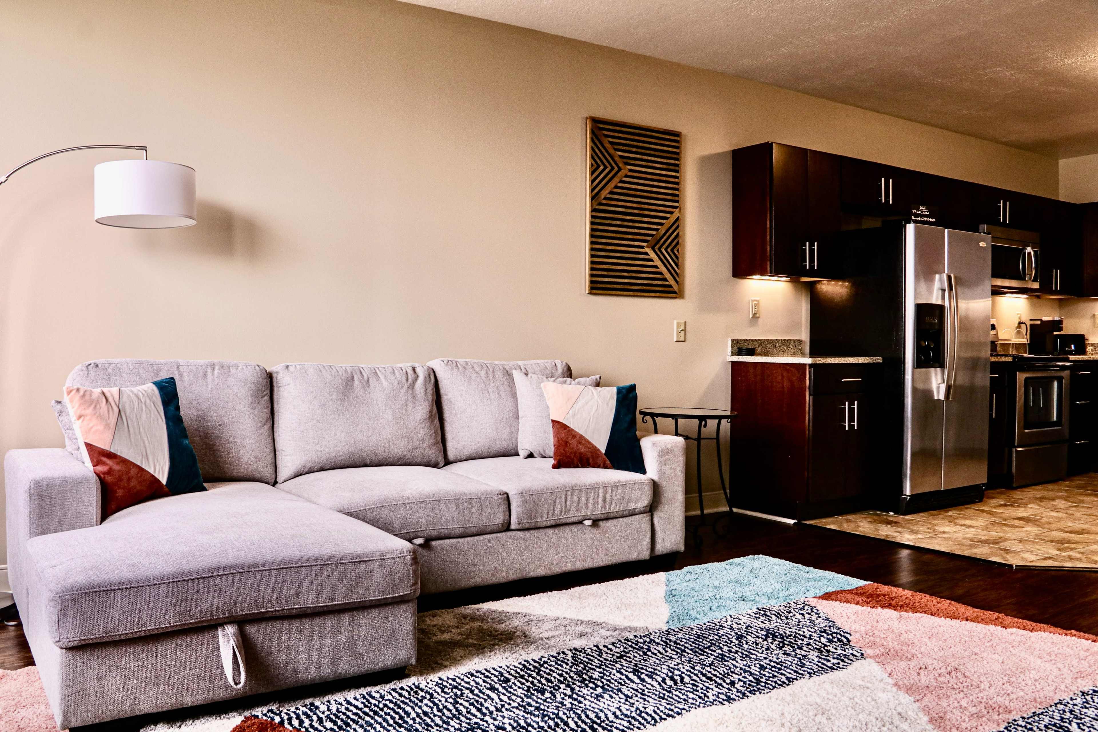The image shows a modern living room featuring a gray sectional sofa, a multi-colored area rug, and a kitchen with dark wooden cabinets and stainless steel appliances.