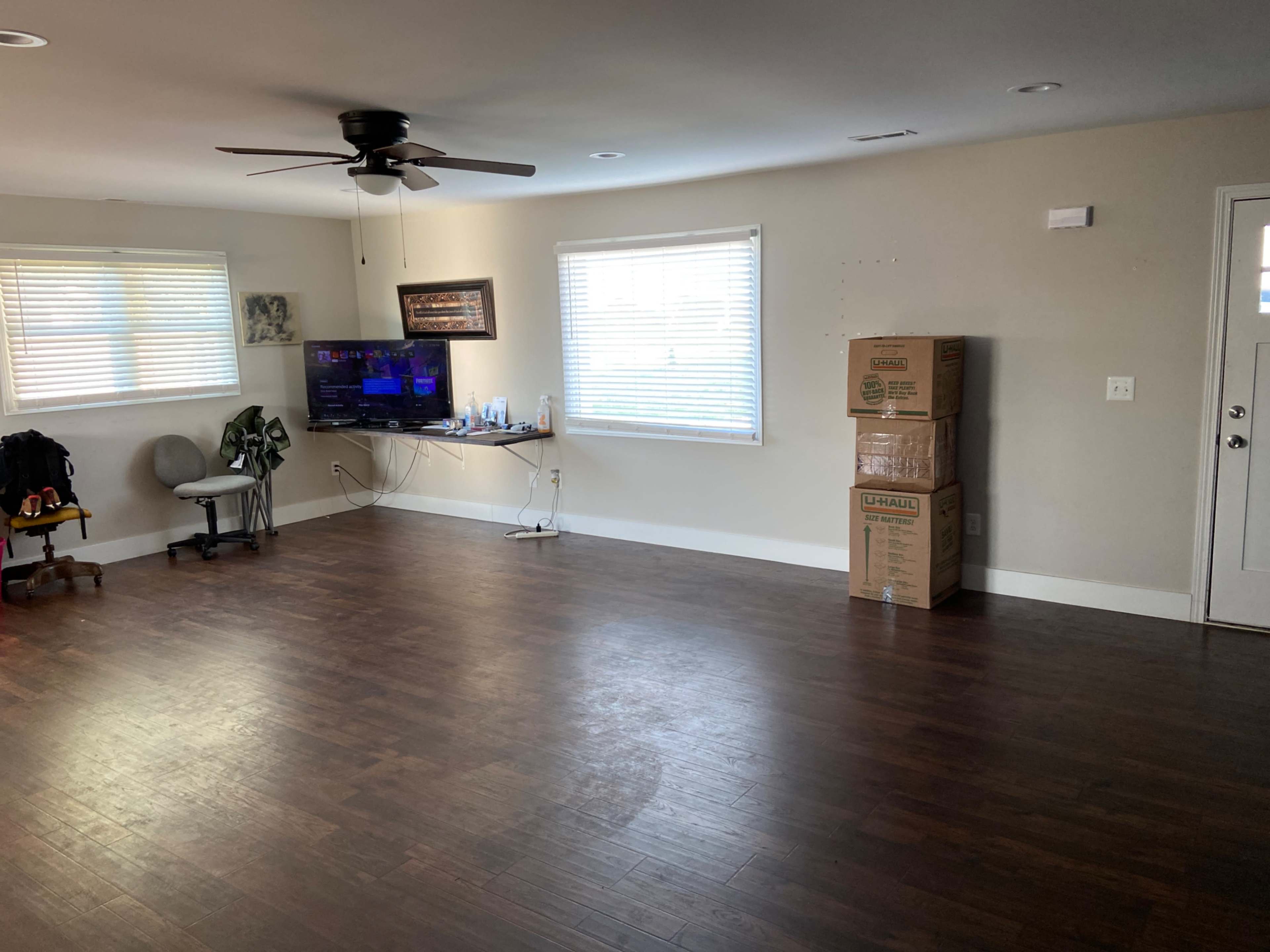 A sparsely furnished living room features a ceiling fan, a wall-mounted television, and a stack of cardboard boxes near the door.