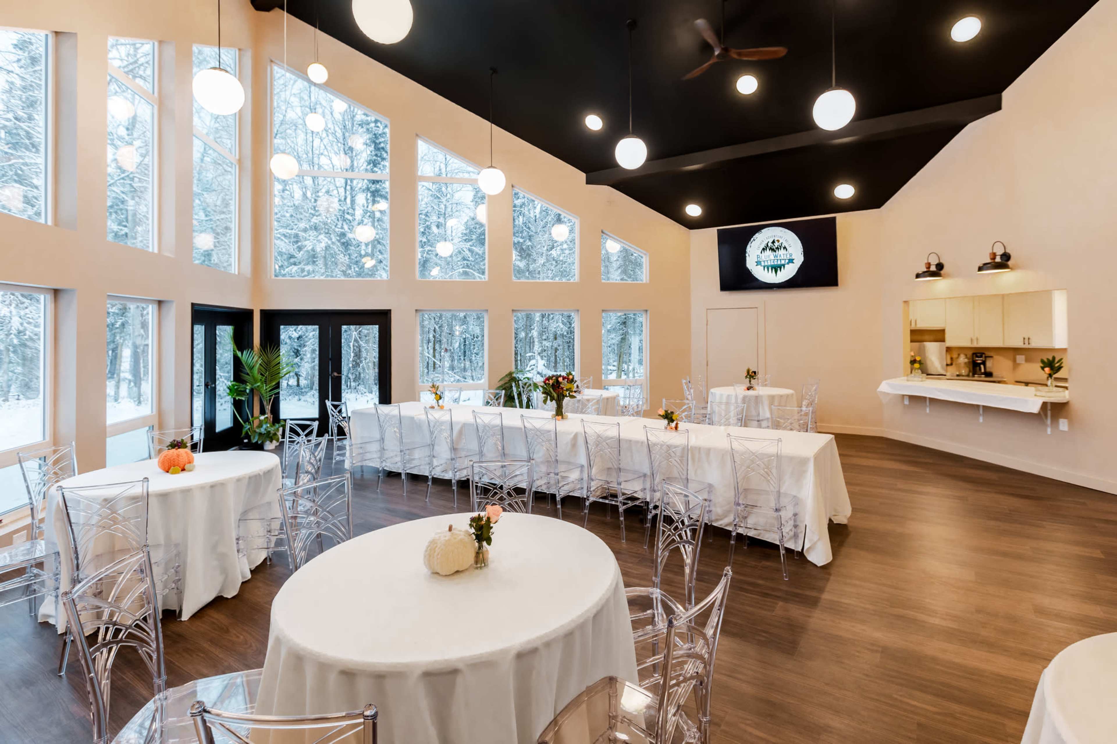 The image shows a spacious indoor venue with large windows overlooking a snowy landscape, featuring tables covered with white tablecloths and modern, transparent chairs.