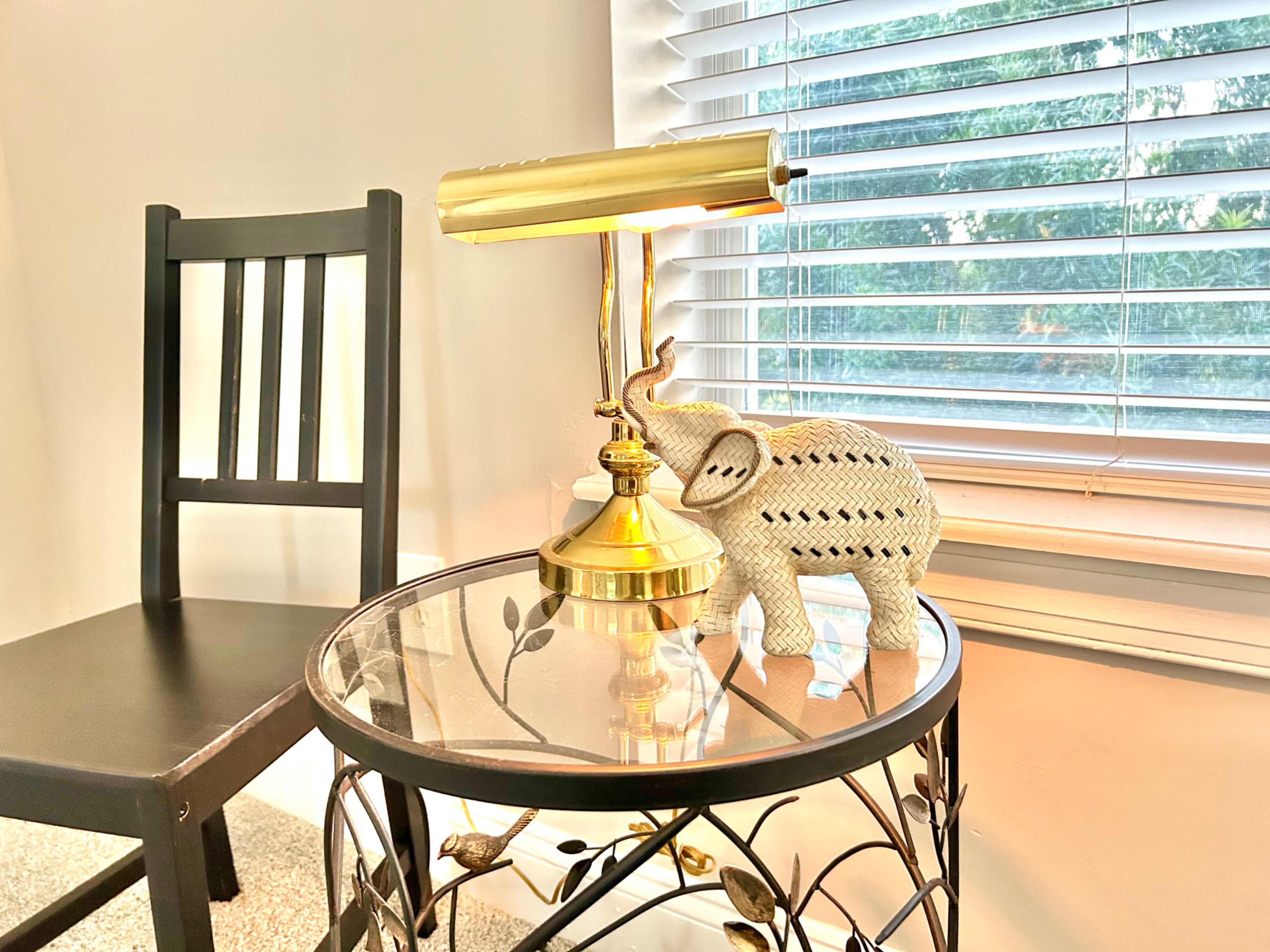 A black chair sits next to a round table with a glass top, featuring a decorative elephant and a gold lamp beside a window with blinds.