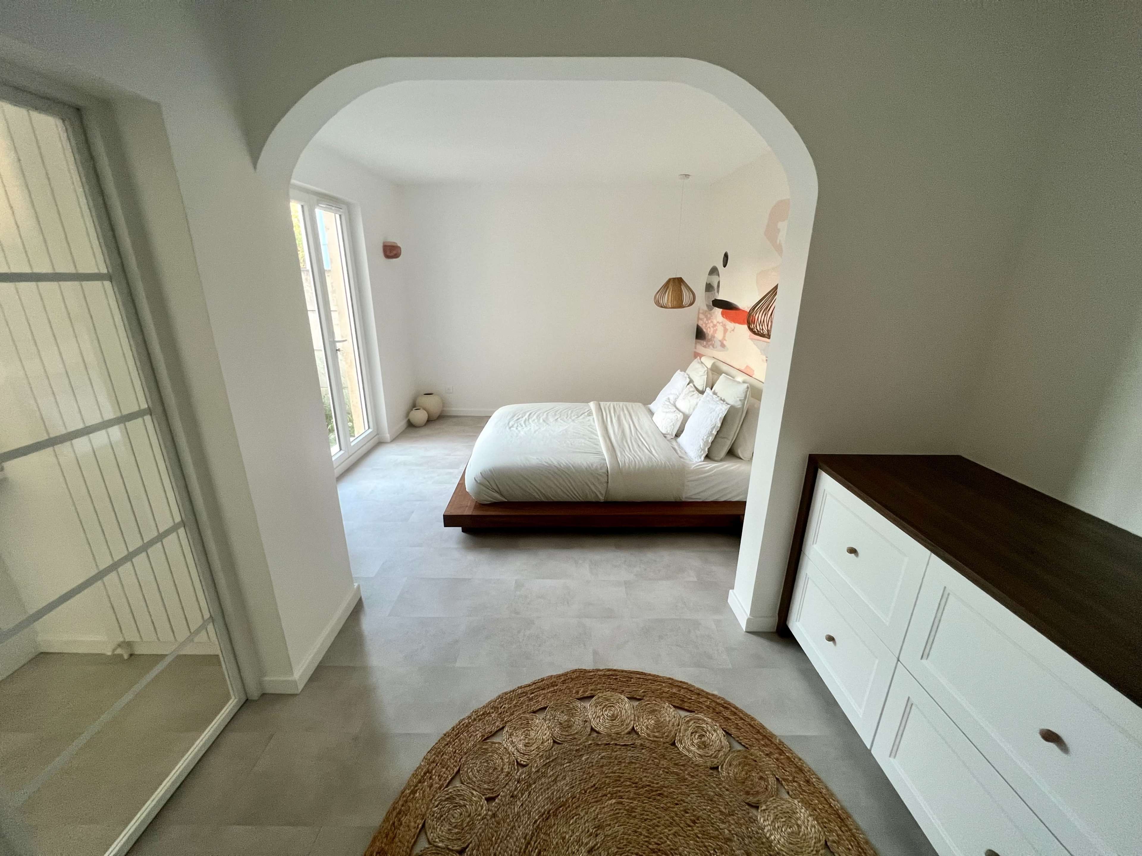 A minimalistic bedroom features a wooden bed, light-colored walls, and large windows with natural light.