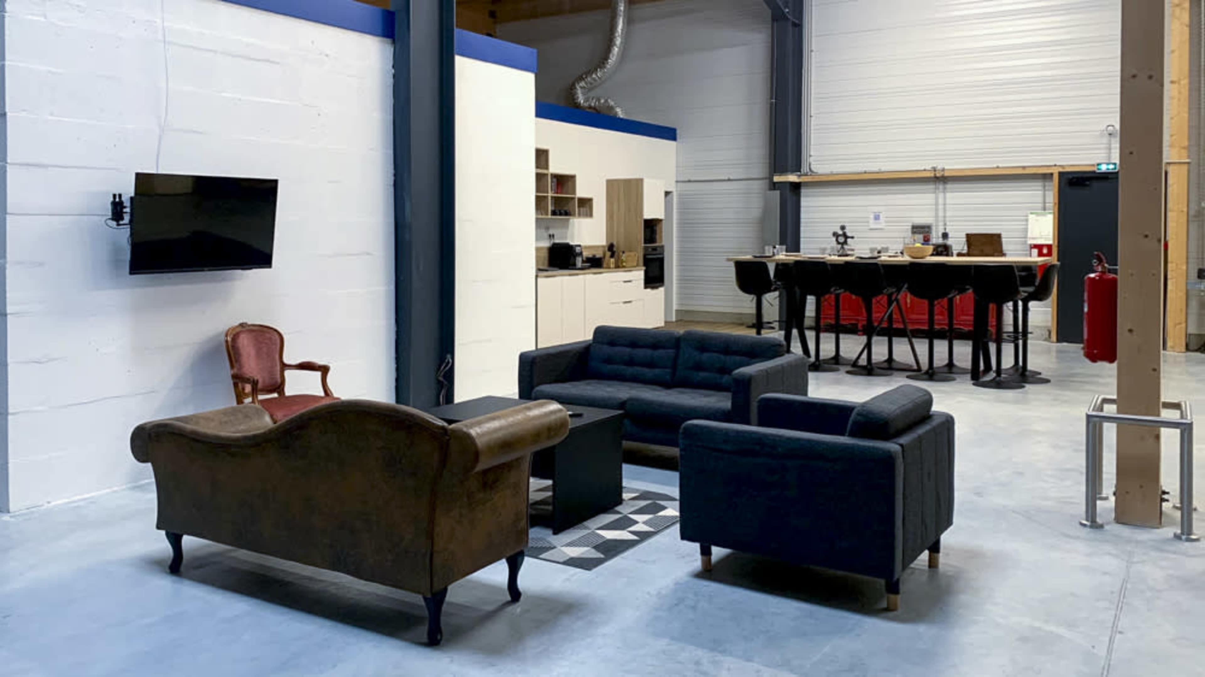The image shows a modern open space featuring a seating area with a brown leather sofa, two dark sofas, and a black coffee table, alongside a kitchen area with a bar table and chairs.