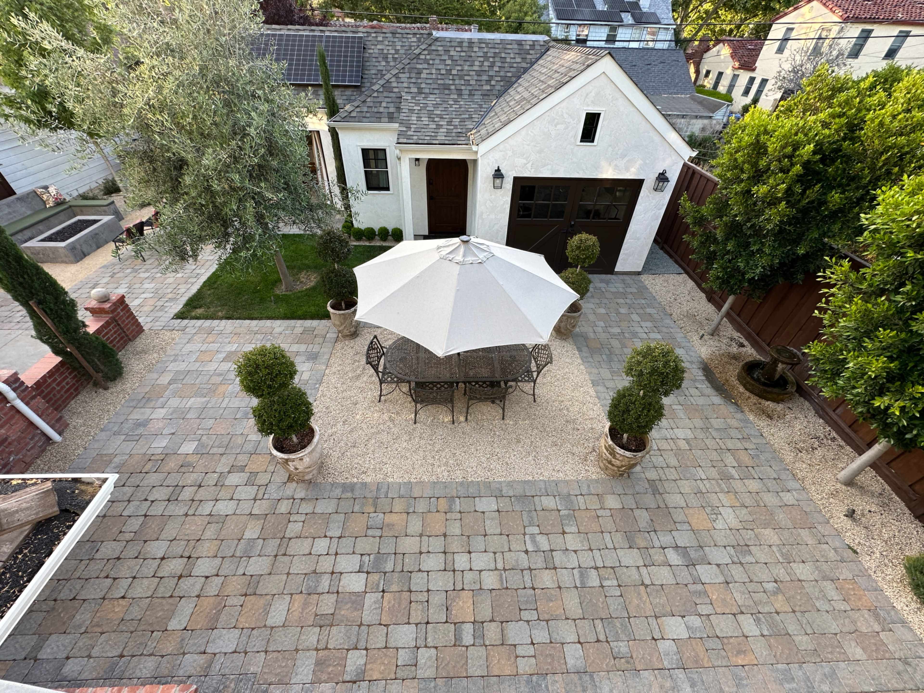 The image shows a well-designed patio area featuring a round table with chairs under a large umbrella, surrounded by decorative shrubs and a gravel pathway.