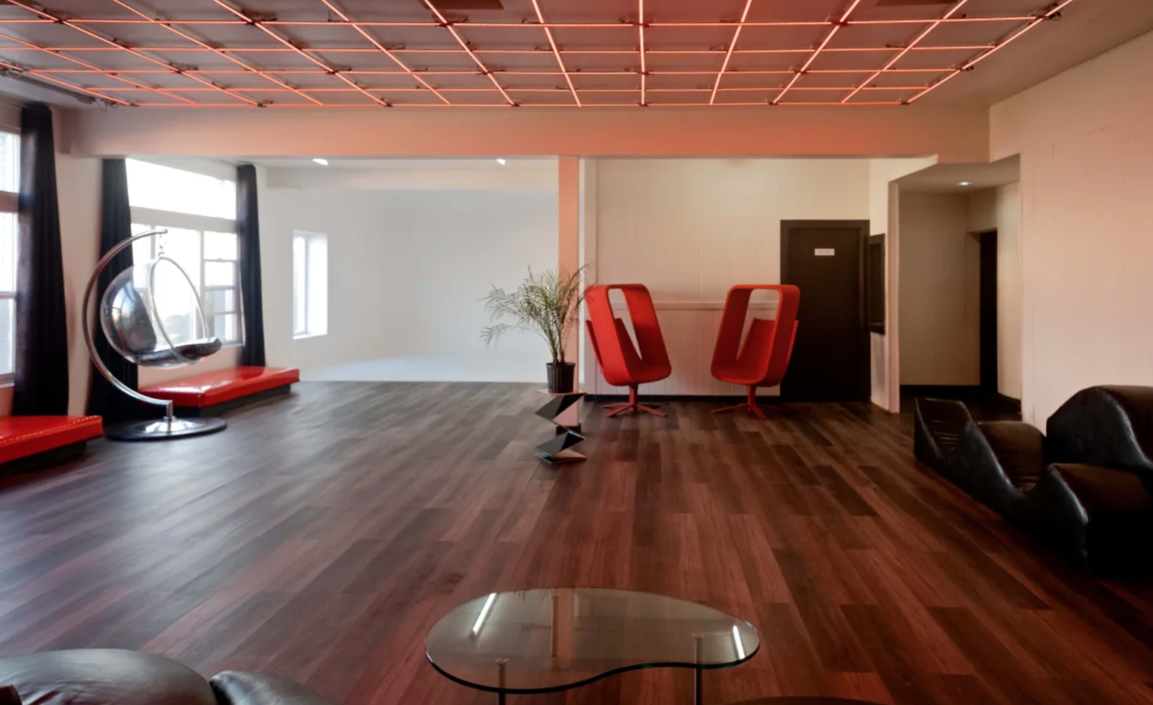 A spacious, modern room with a wooden floor, featuring red seating areas, two red chairs, a hanging chair, and a geometric black sculpture.