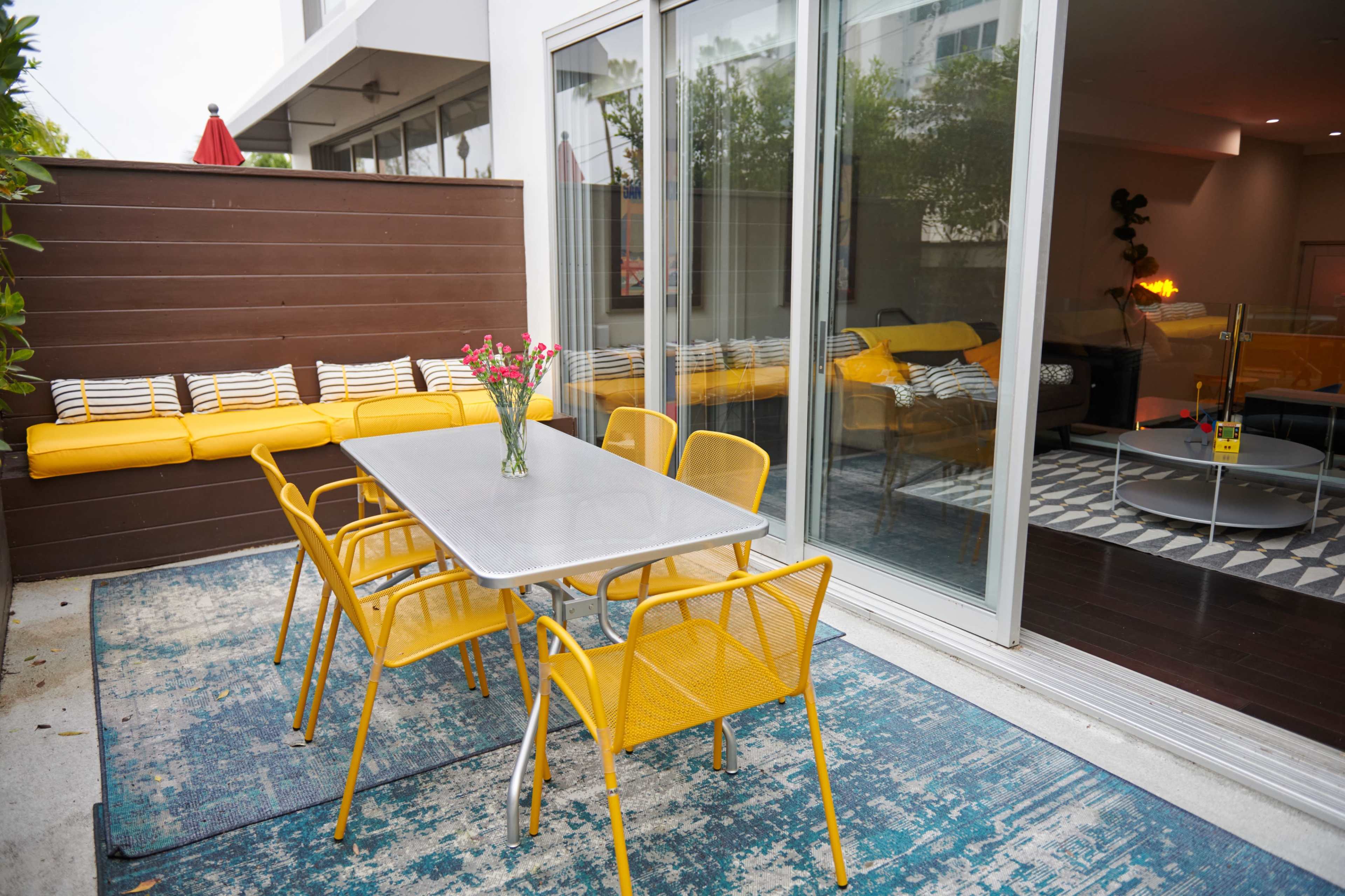 A metal table with yellow chairs is set on a blue rug outside a modern living space, with a yellow cushioned bench visible in the background.