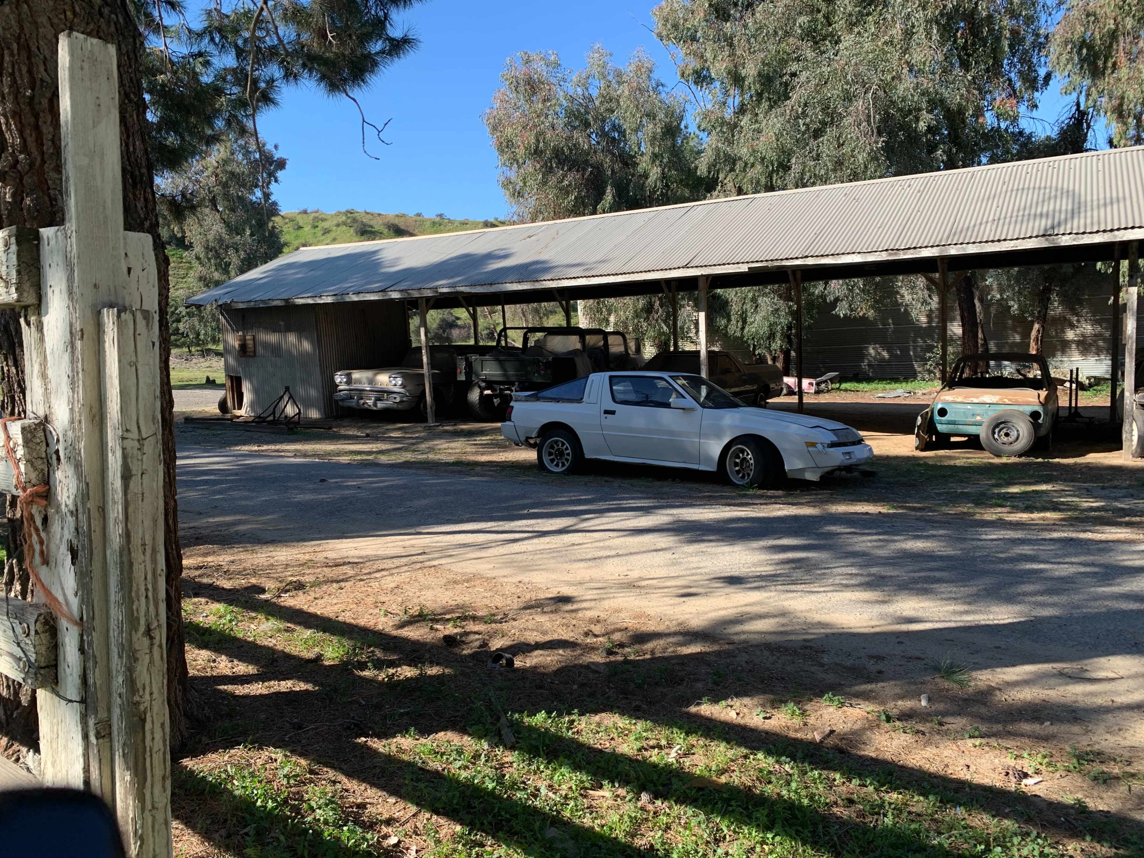 Several parked vehicles, including a white car, under a metal roof shelter surrounded by trees and open land.
