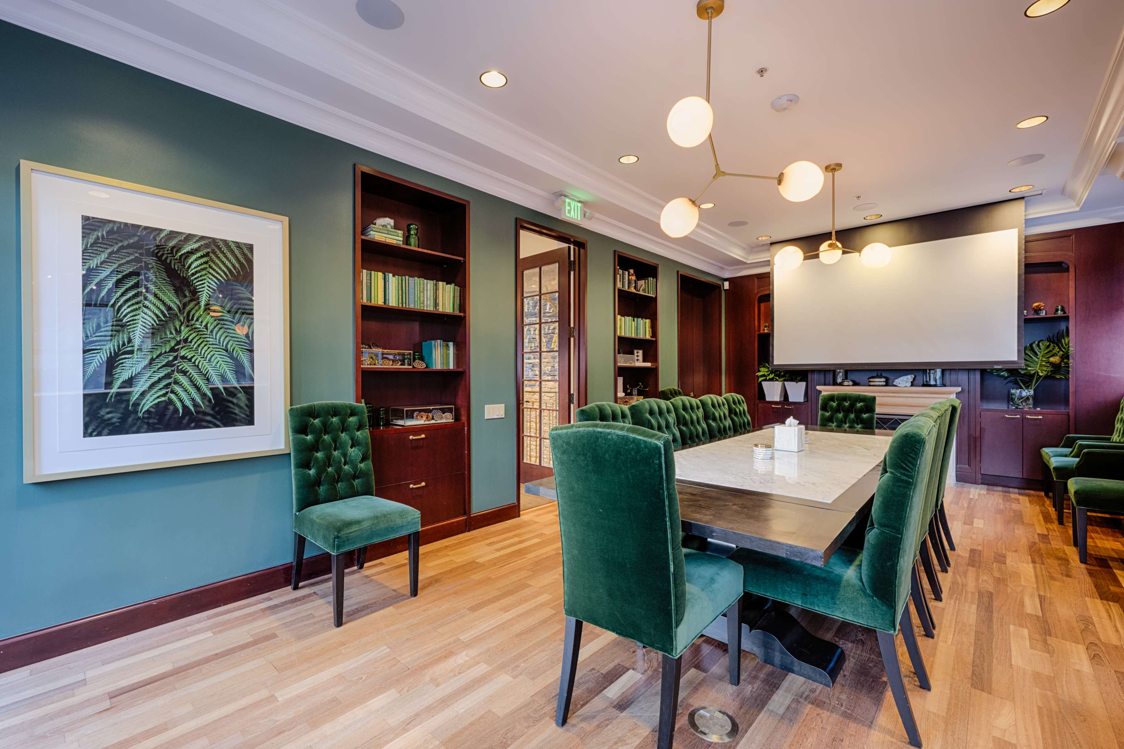 A conference room features a large table surrounded by green upholstered chairs, with shelves of books and a projector screen on the wall.