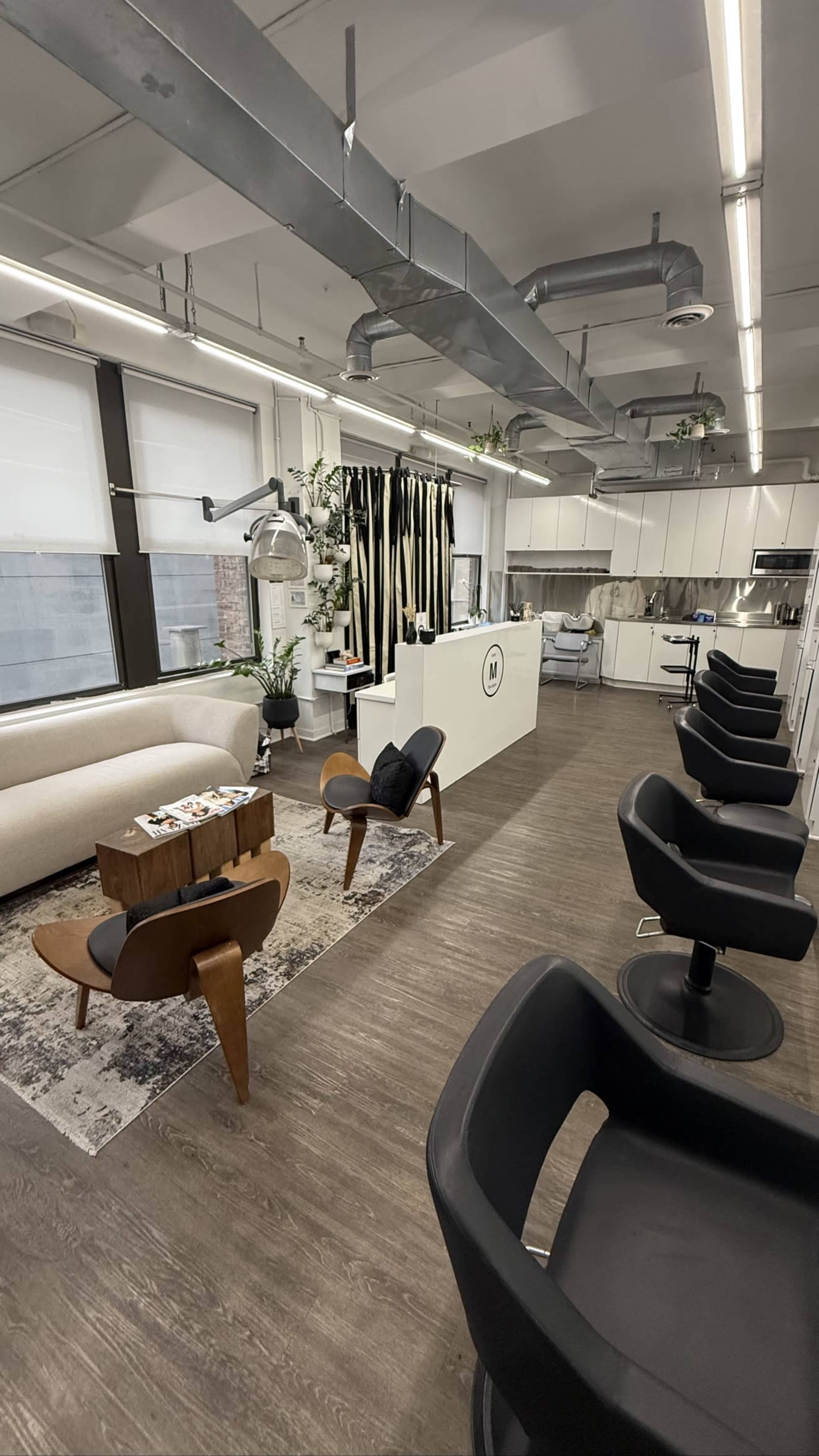 The image shows a modern, well-lit salon interior featuring black salon chairs, a reception area with a white counter, and large windows with white blinds.