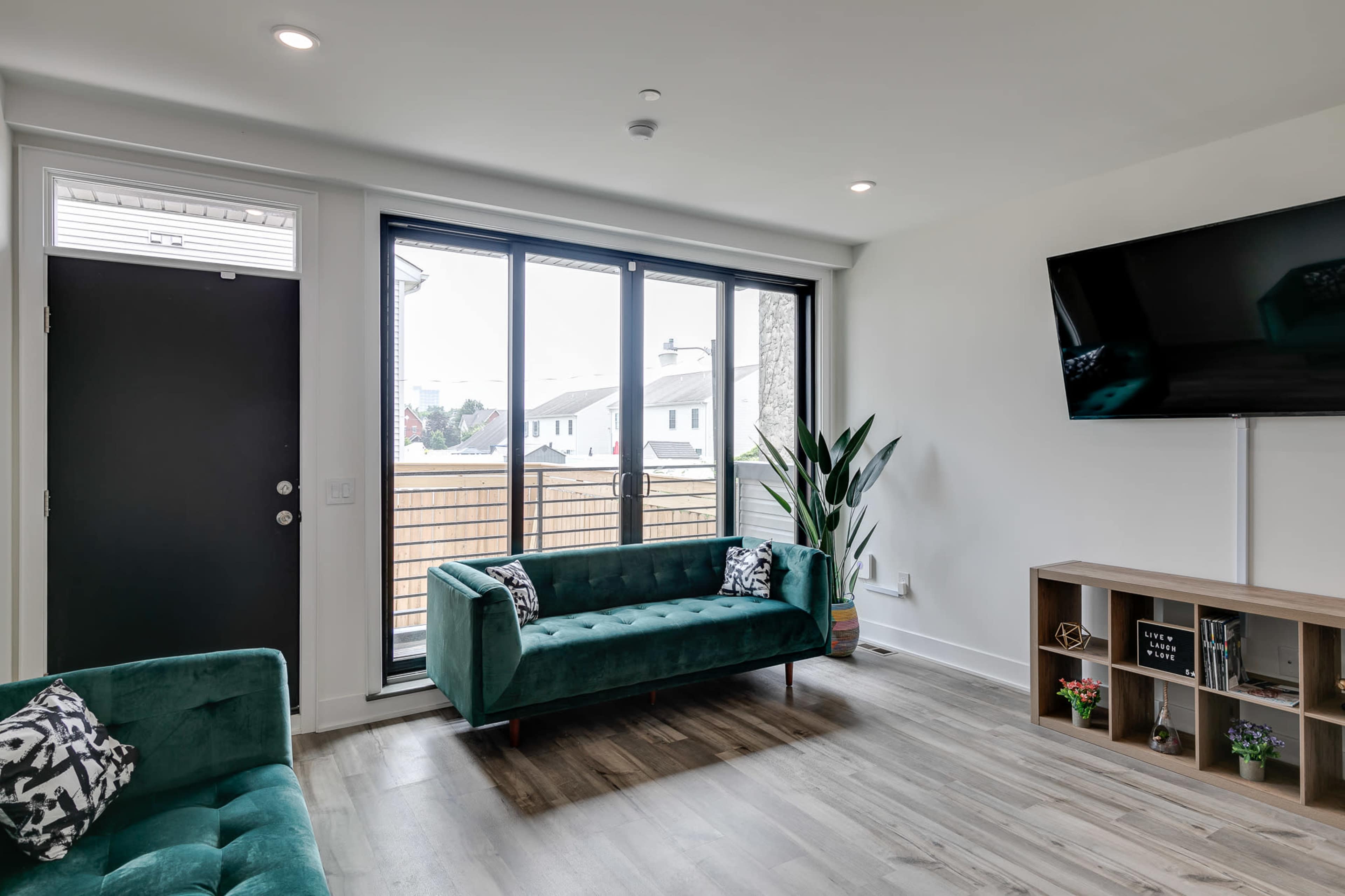 A modern living room with two green sofas, a television mounted on the wall, a wooden shelf with decorative items, and a large window overlooking an outdoor space.