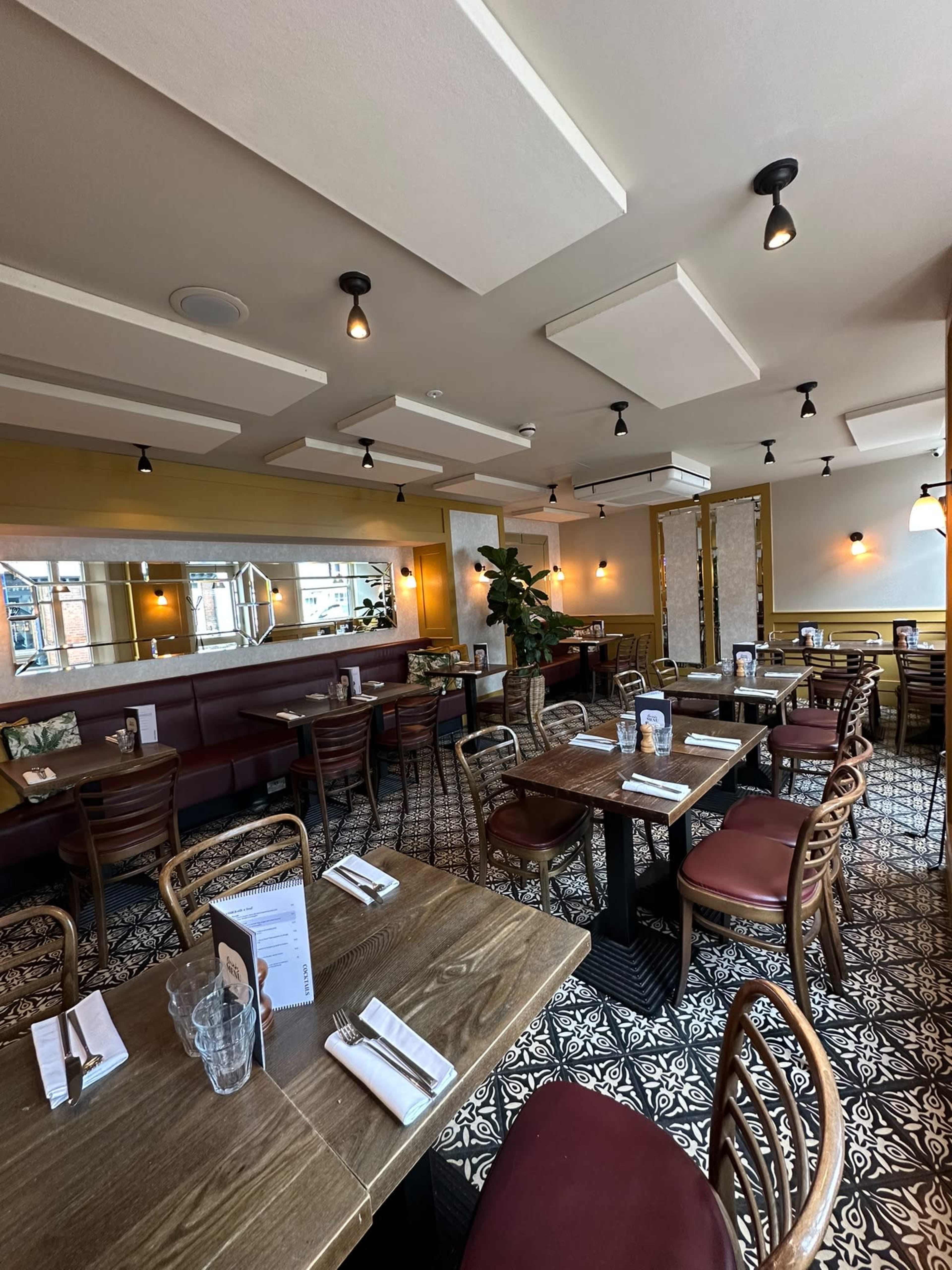 The image shows the interior of a restaurant featuring wooden tables, burgundy chairs, and patterned tile flooring, with a mix of decorative plants and ambient lighting.