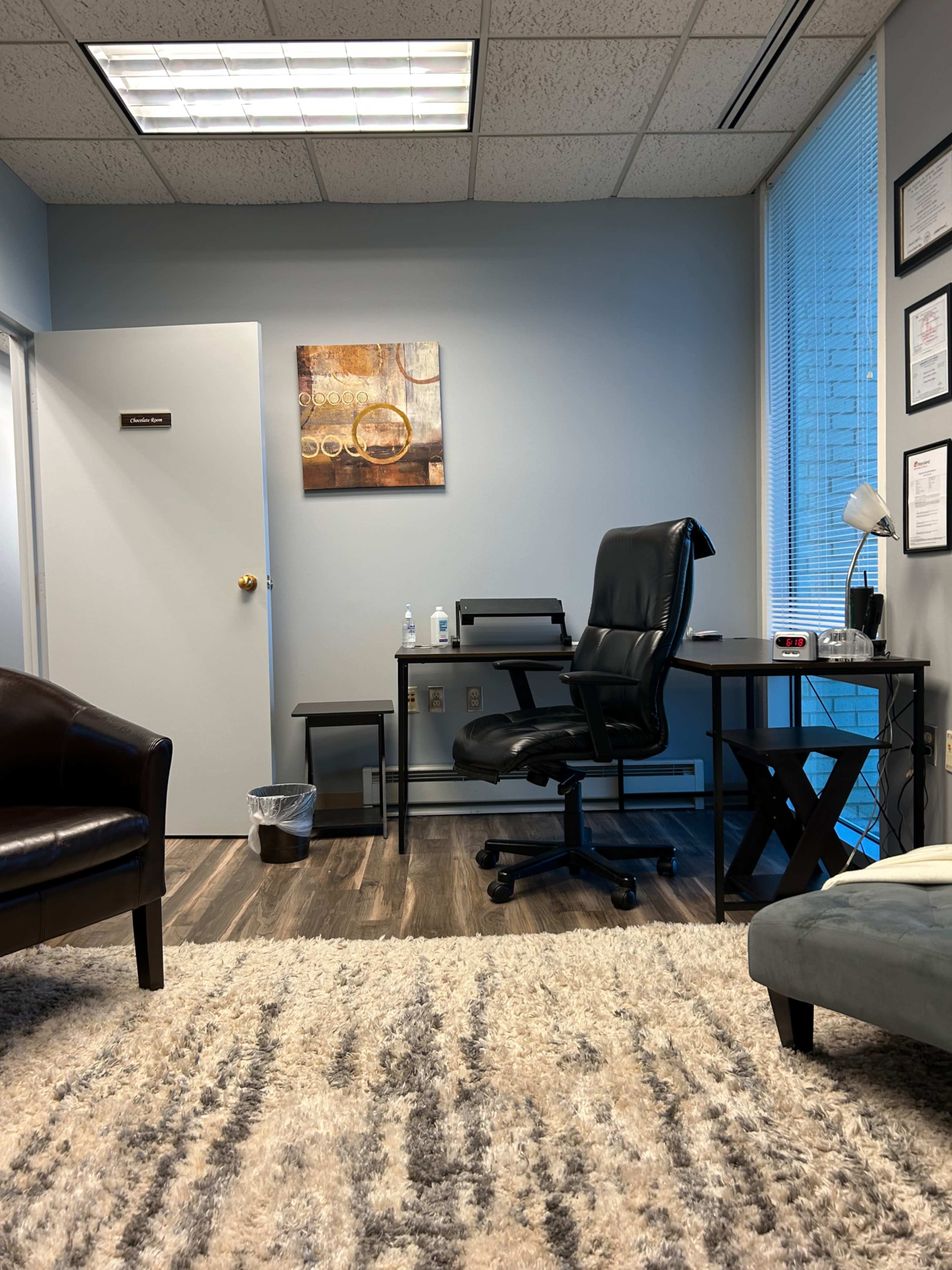 The image shows a neatly arranged office with a desk, chair, and wall art, along with a door and large window.