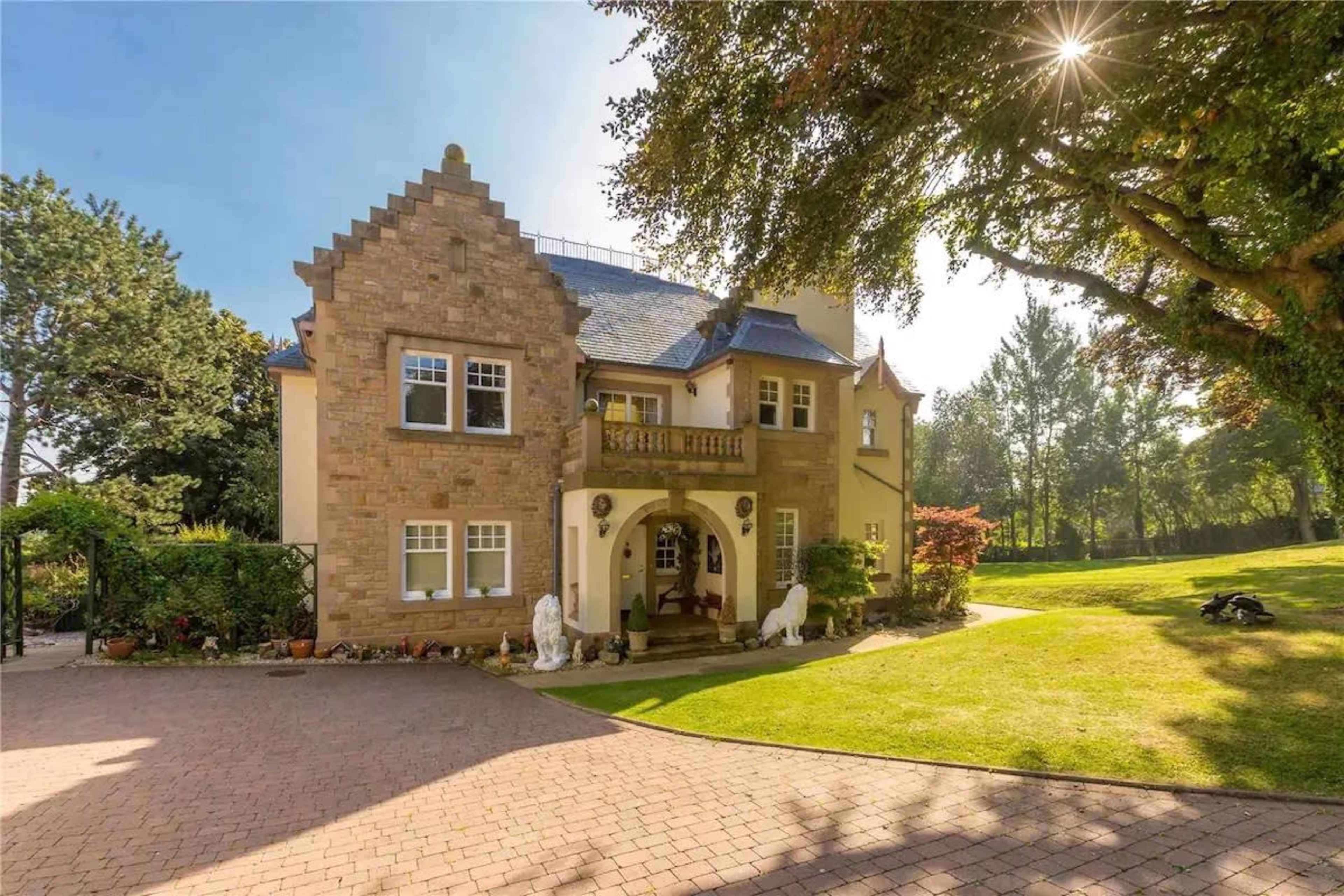 A large stone house with intricate architectural details stands amidst a landscaped yard under a clear sky.
