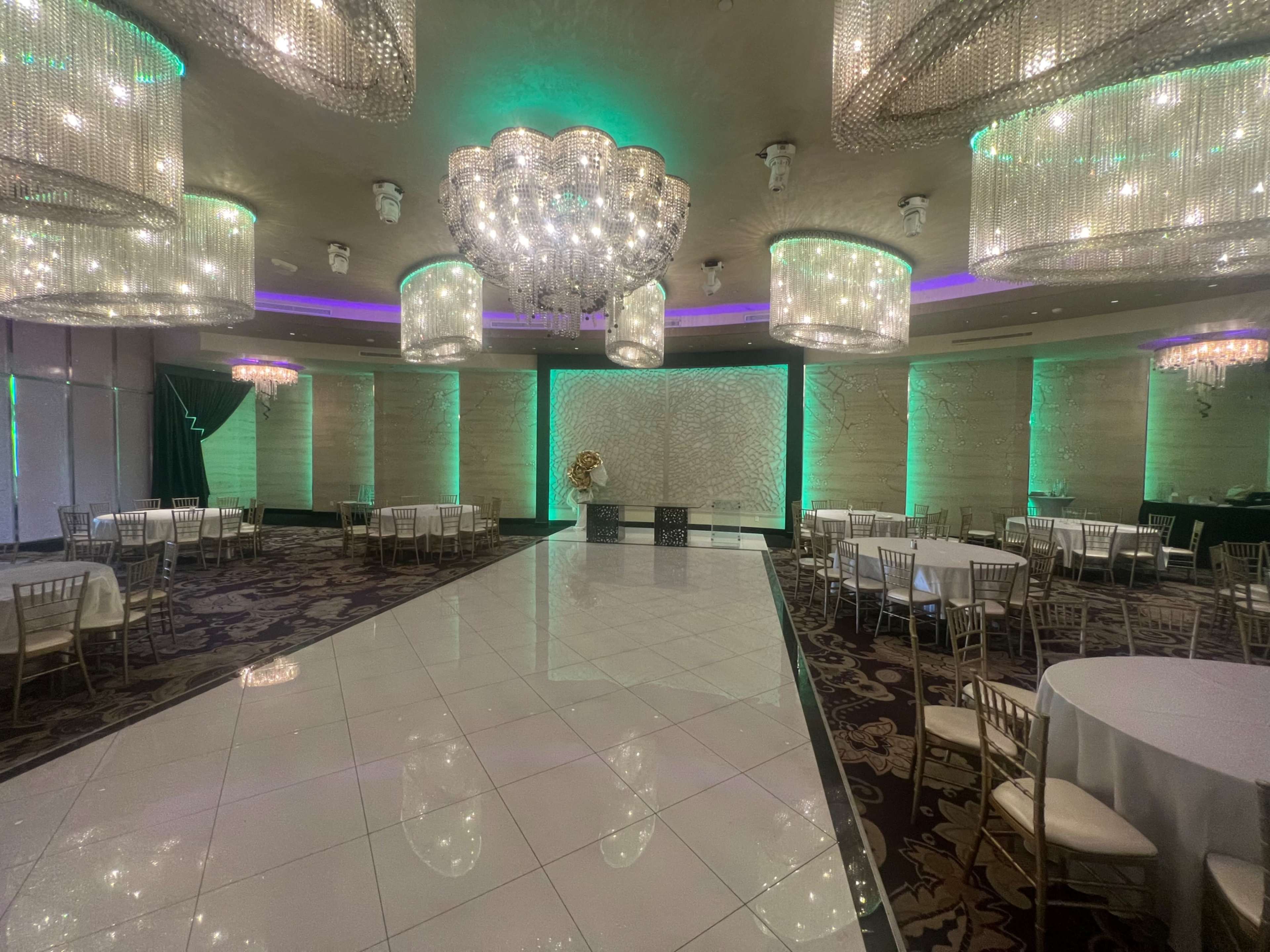 A large event space features white tables and gold chairs under illuminated chandeliers, with a polished dance floor and textured wall panels.