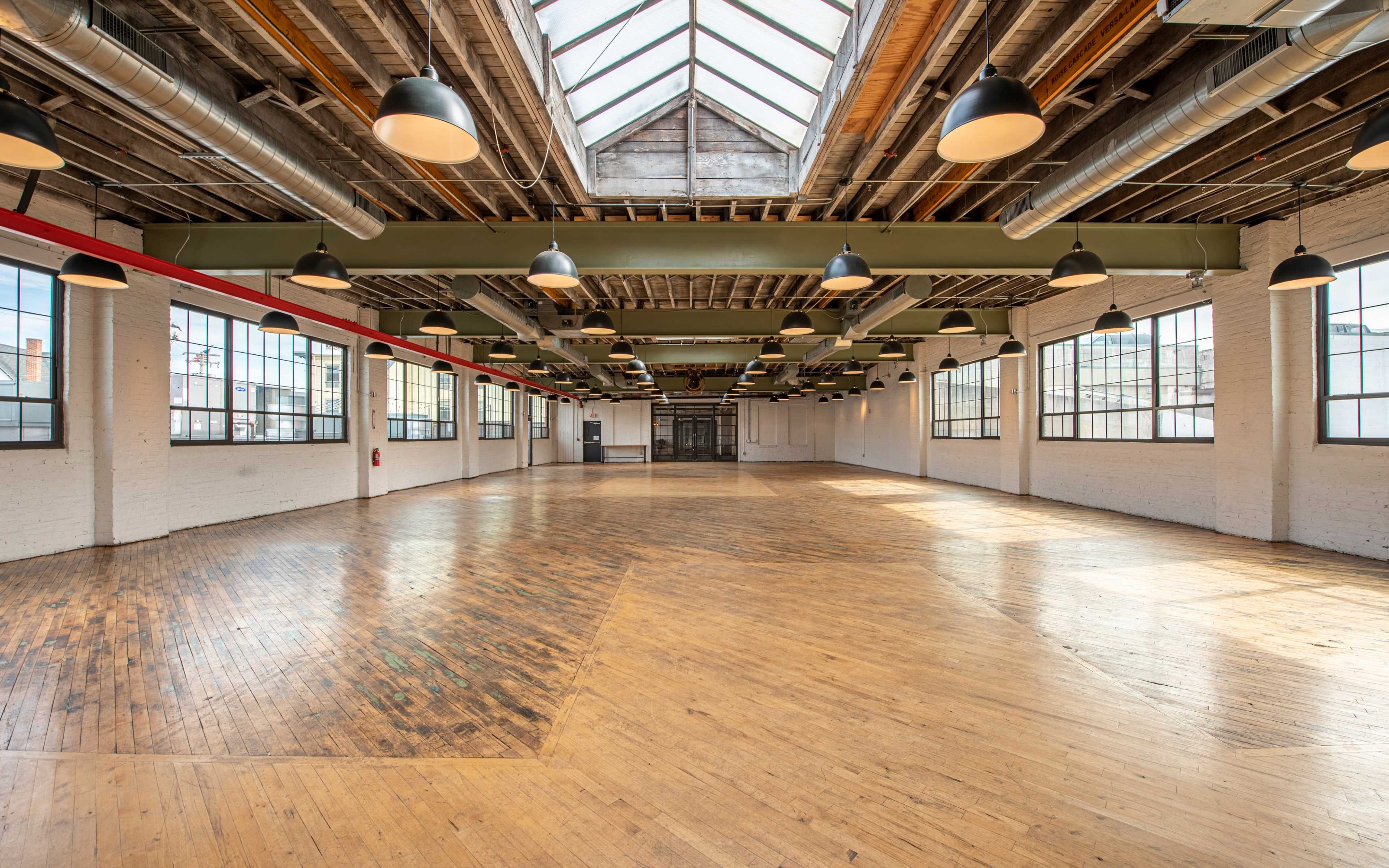 A spacious, empty industrial-style room with wooden floors, large windows, and overhead pendant lights.