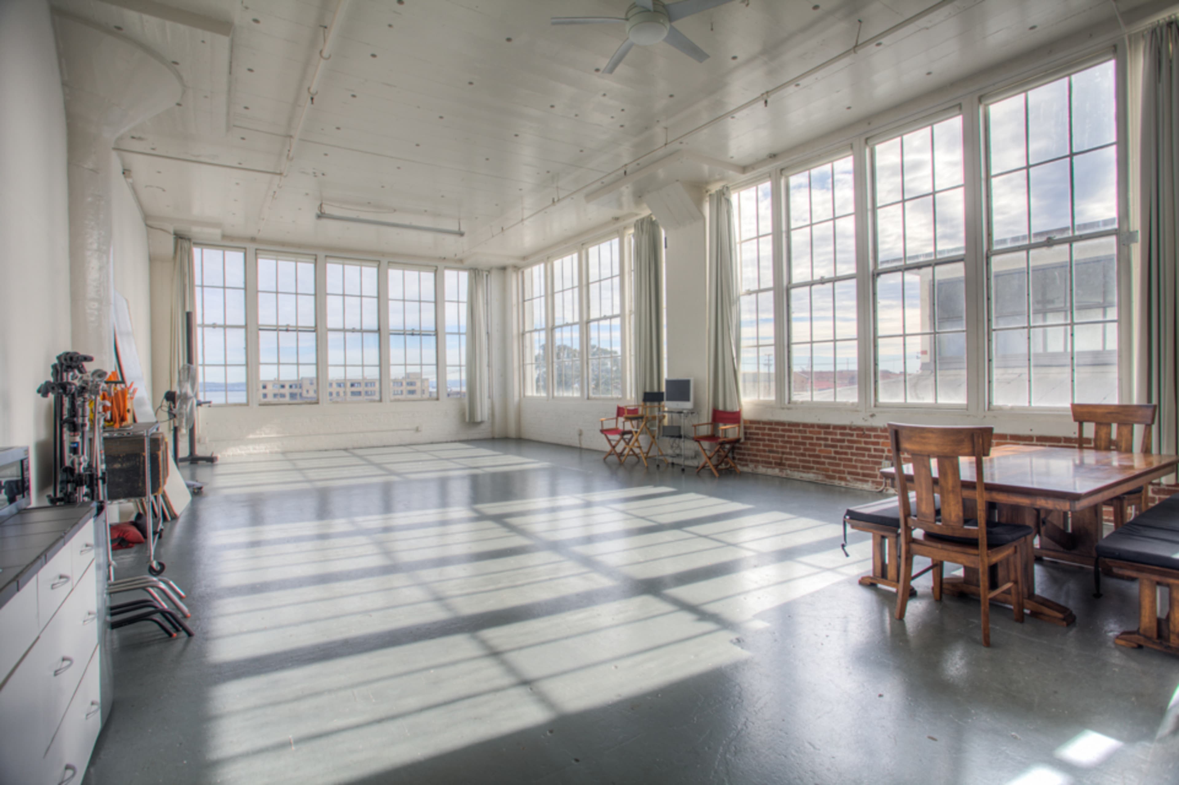 A spacious room with large windows, wooden furniture, and a dining table, illuminated by natural light.