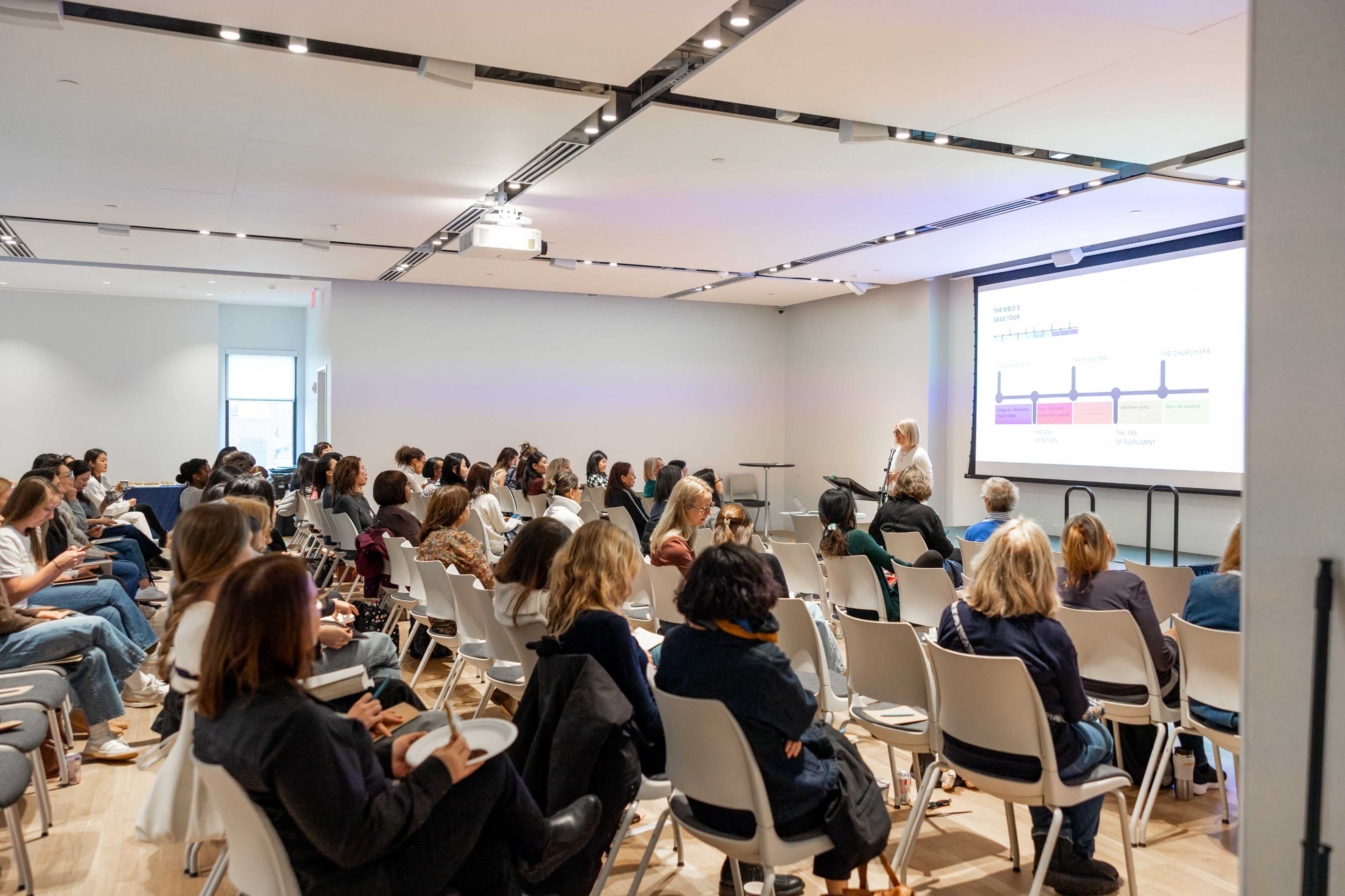 A group of people are seated in a conference room while an individual presents information using a projected screen.