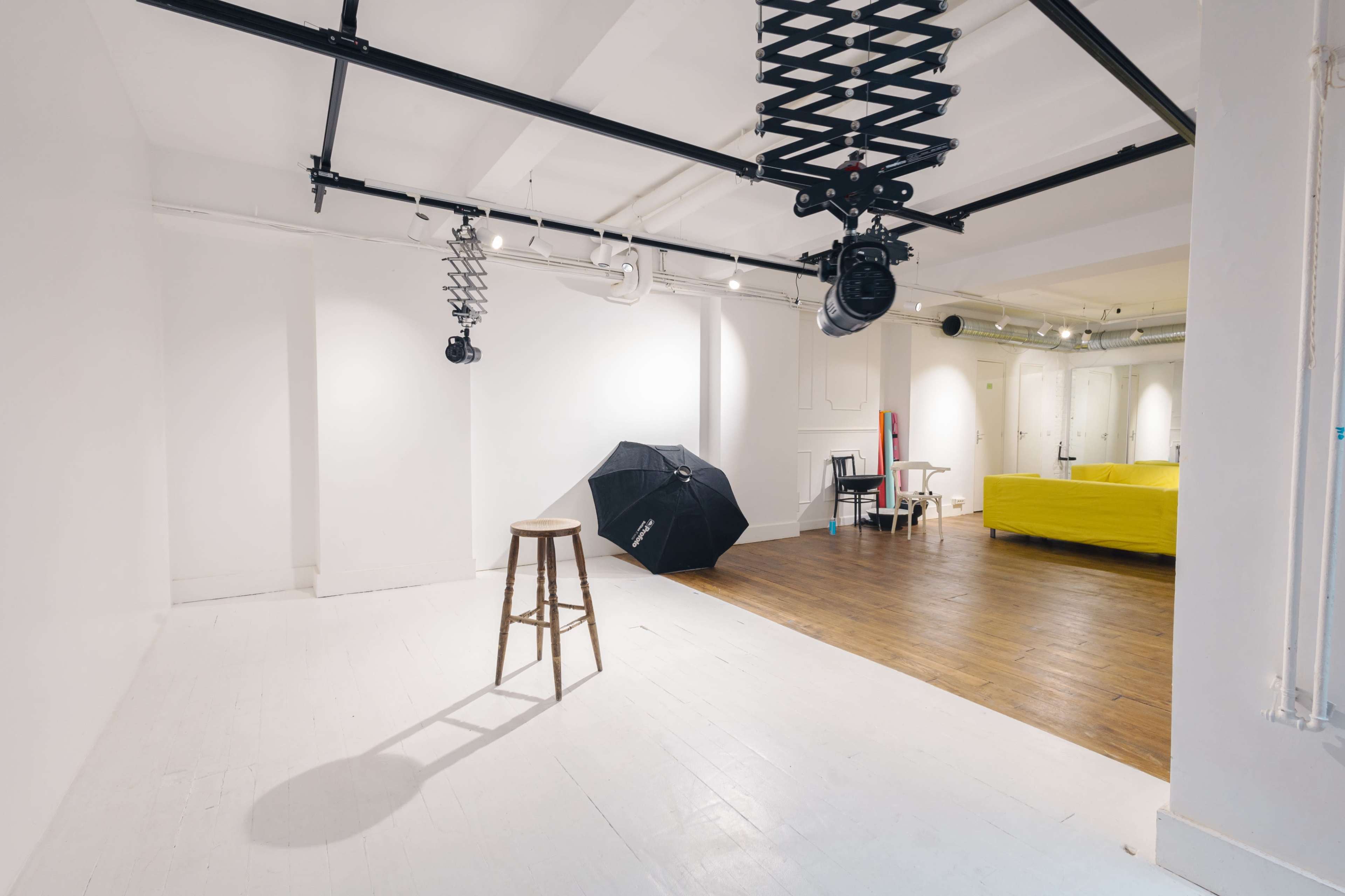 A spacious, minimalist studio with a wooden stool, a black umbrella, and a yellow couch in the background.
