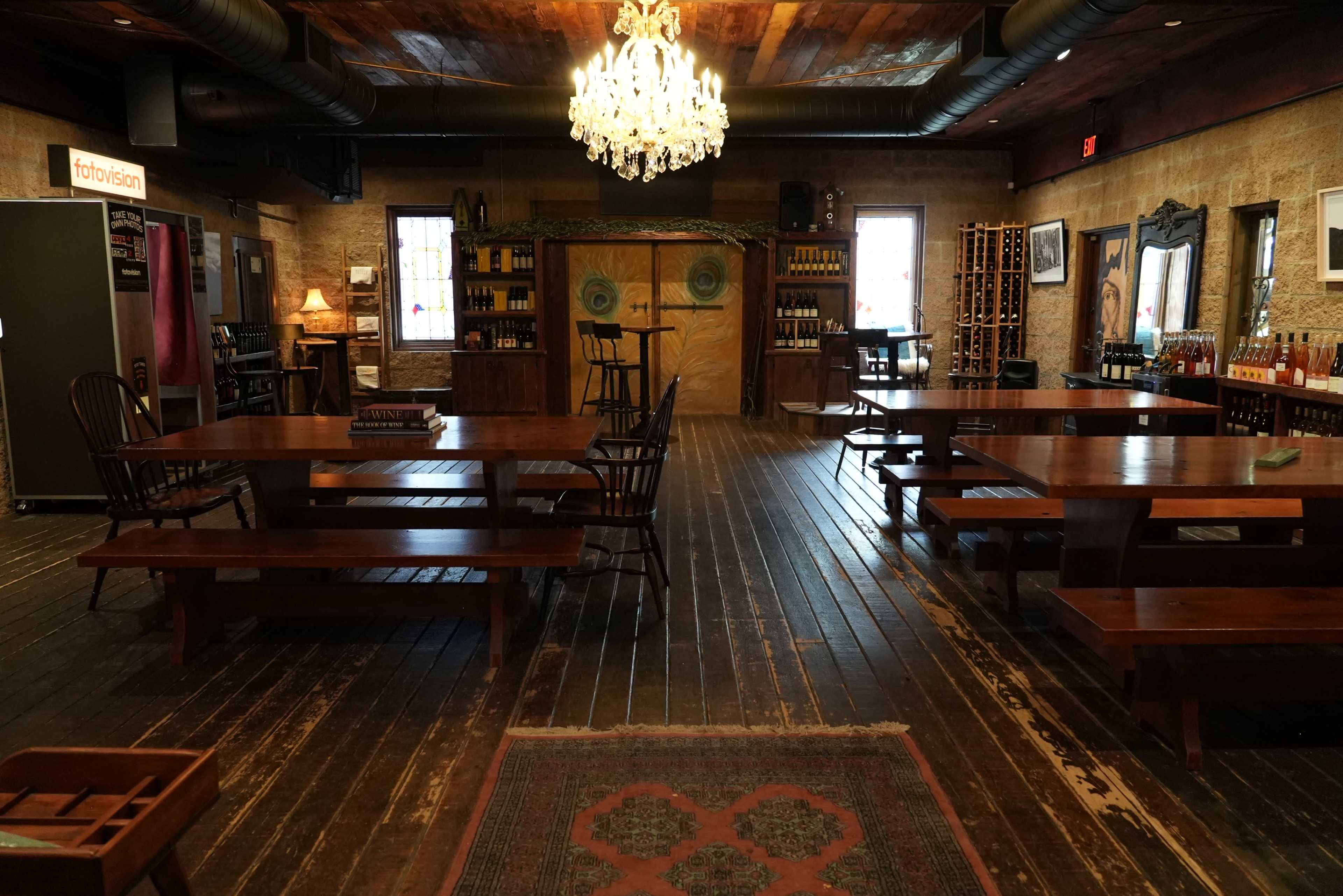 The image shows a spacious wooden interior of a restaurant or tasting room with multiple long tables, decorative lighting, and shelves lined with bottles.