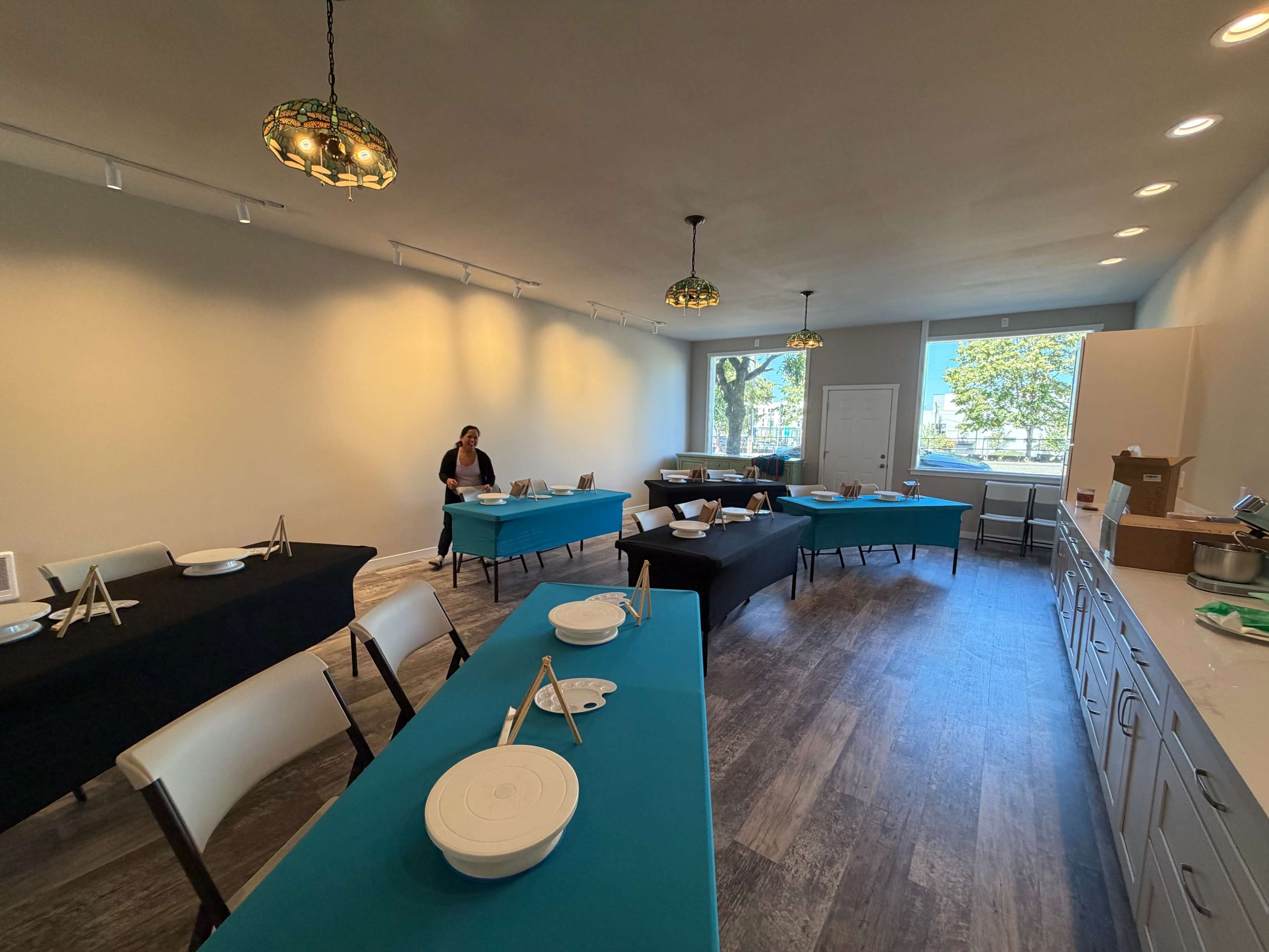 The image shows a spacious room with several tables covered in turquoise tablecloths and a person seated at one of the tables, while a kitchen area is visible at the far end.