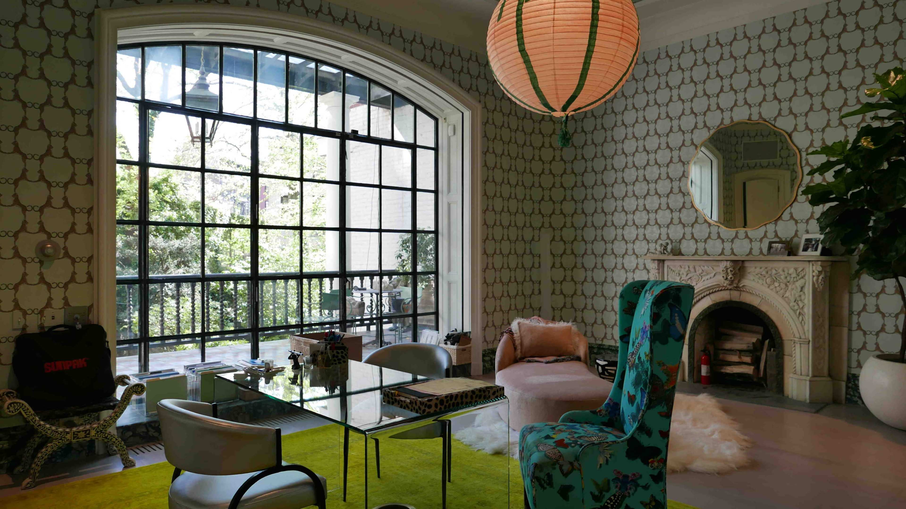 A modern living room features a glass table, a patterned wall, a large window overlooking greenery, and eclectic seating arrangements.