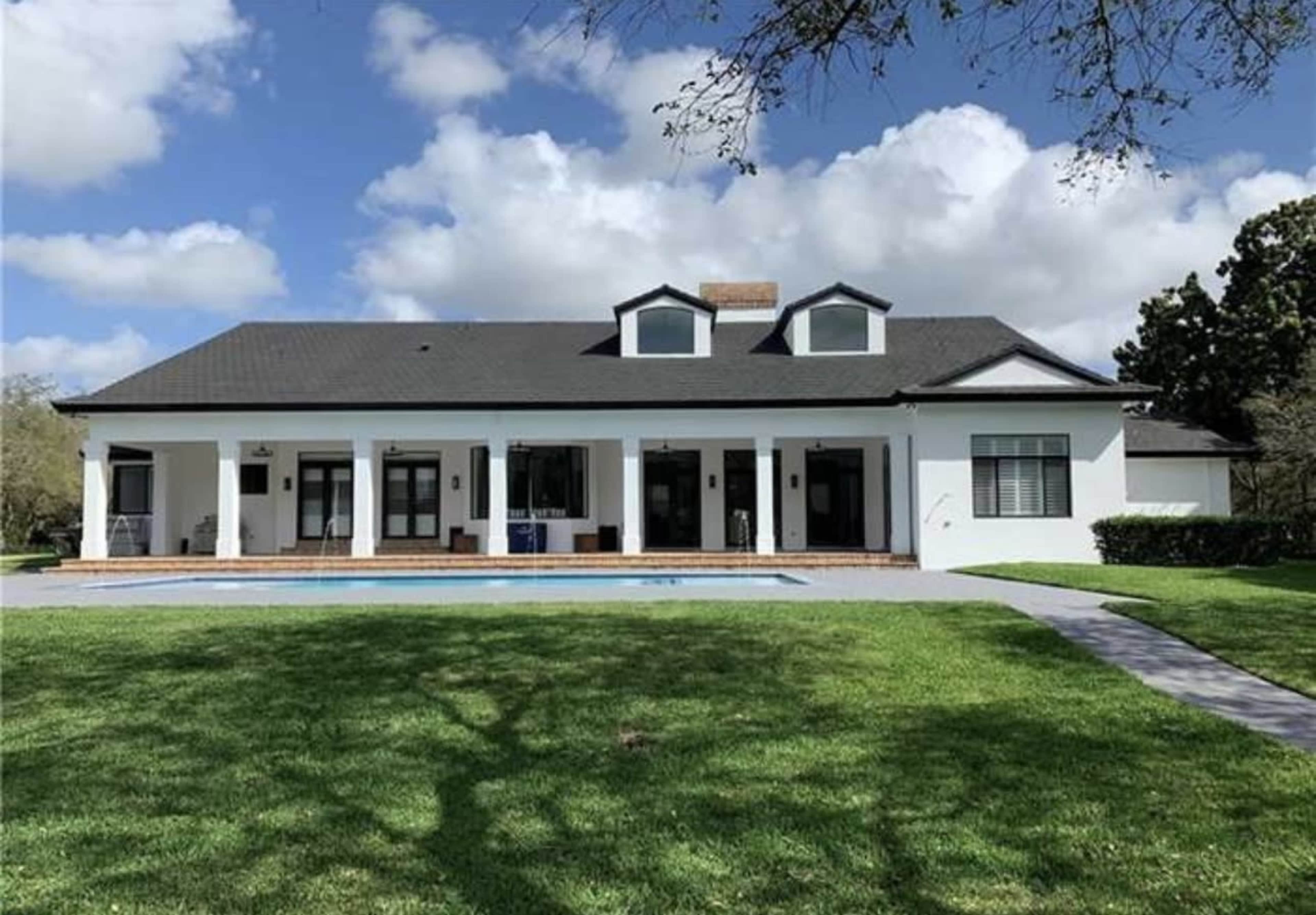 A single-story white house with a black roof and a pool in the foreground is set on a grassy lawn under a partly cloudy sky.