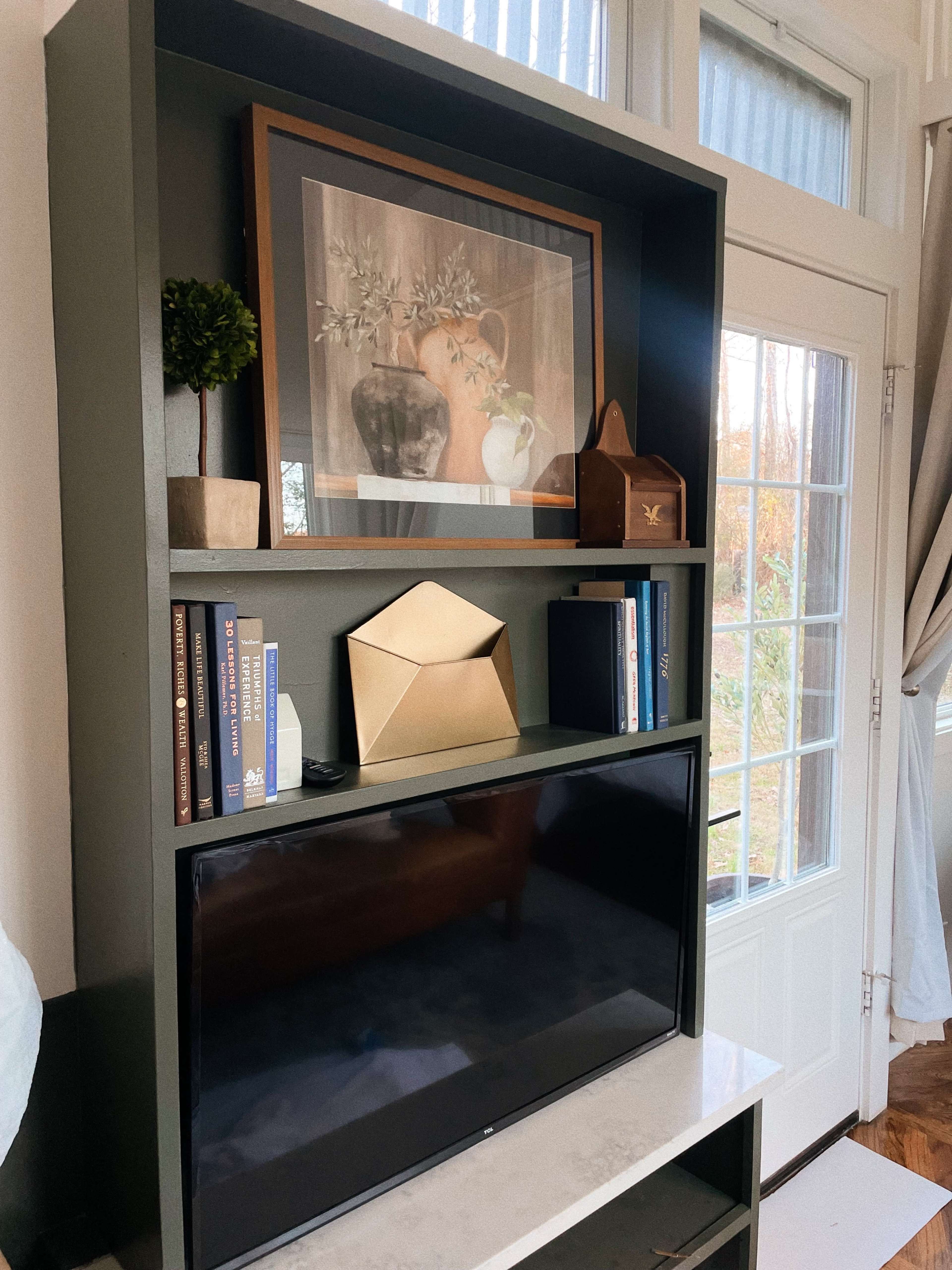 A modern bookshelf containing a framed artwork, decorative items, and a television, positioned next to a window with natural light.