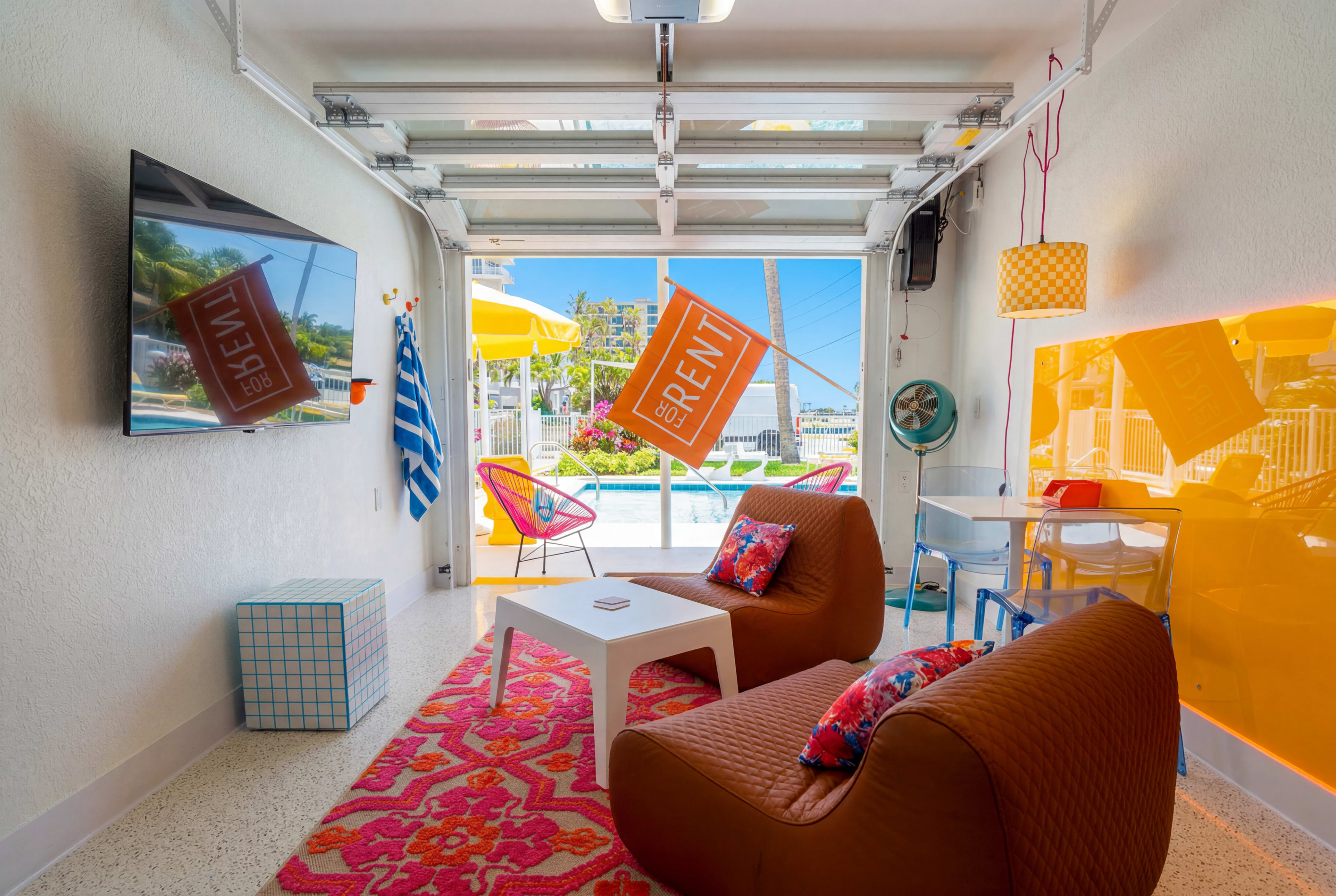 The image shows a colorful lounge area with two modern chairs, a small table, and a television, all set against a backdrop of large open doors leading to a sunny outdoor space.
