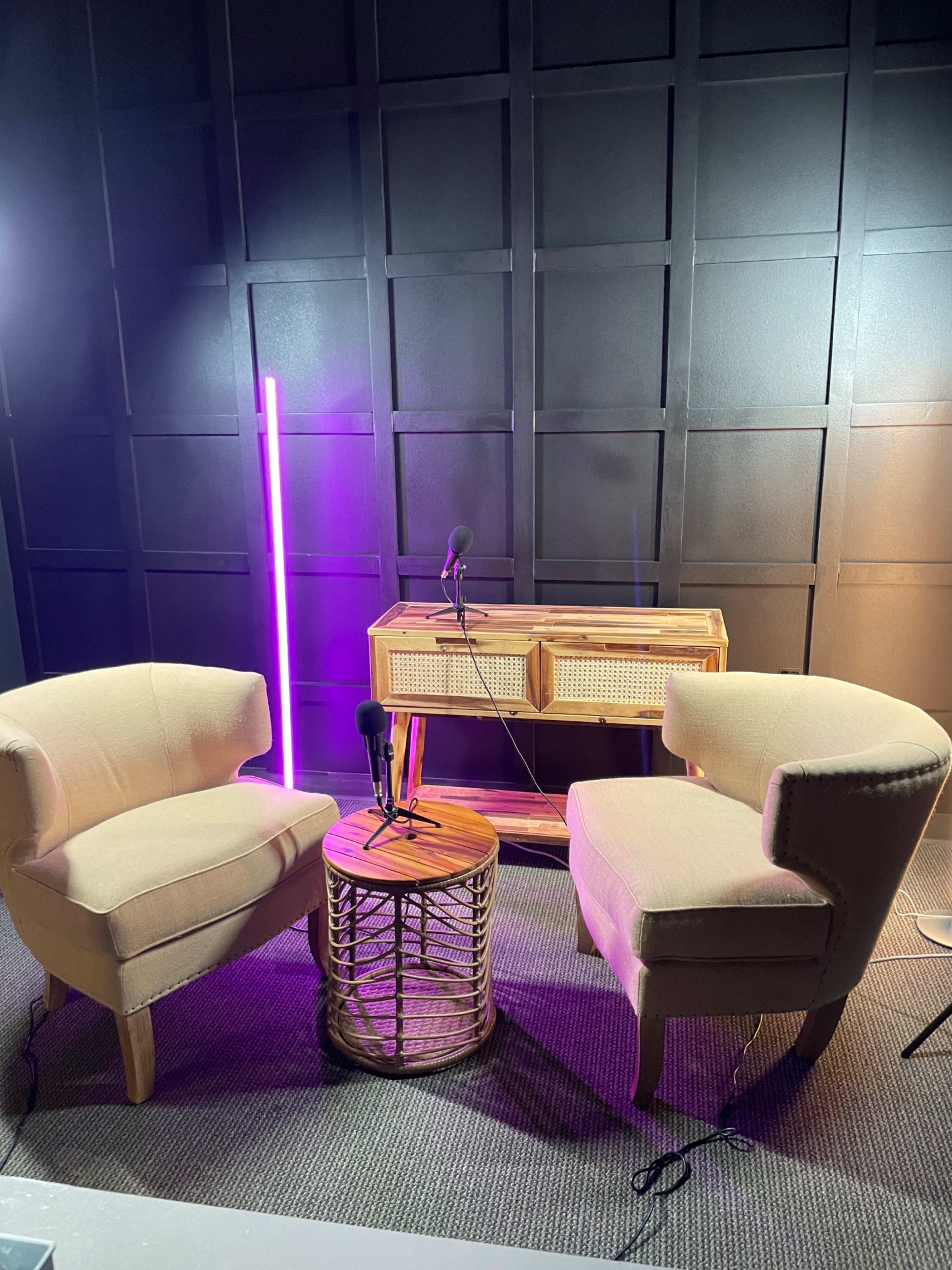 A small studio setup with two upholstered chairs, a round table, and microphones positioned between them against a textured dark wall.