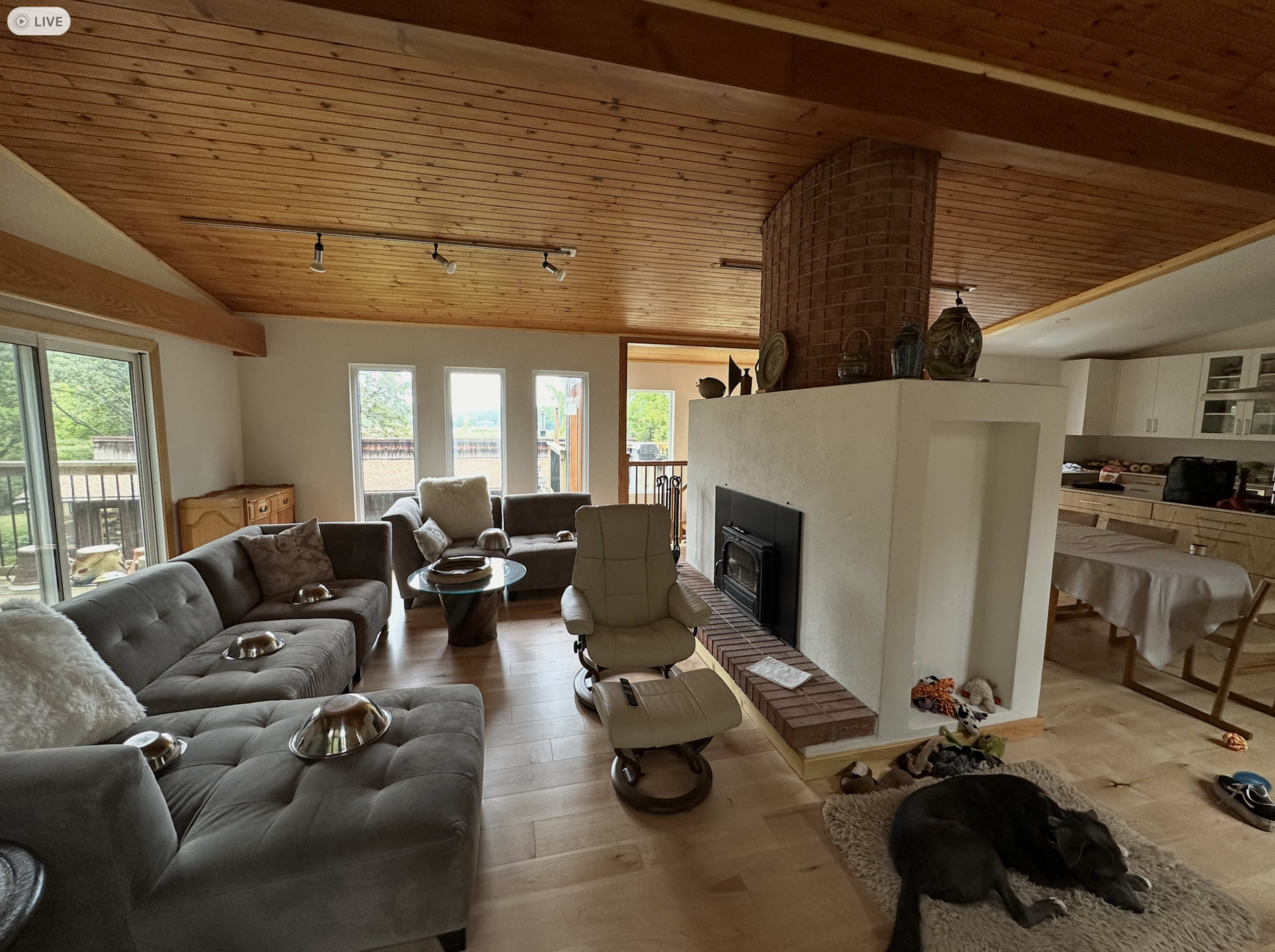 The image shows a spacious living area with a light wood ceiling, a central fireplace, comfortable seating, and large windows that open to an outdoor patio.