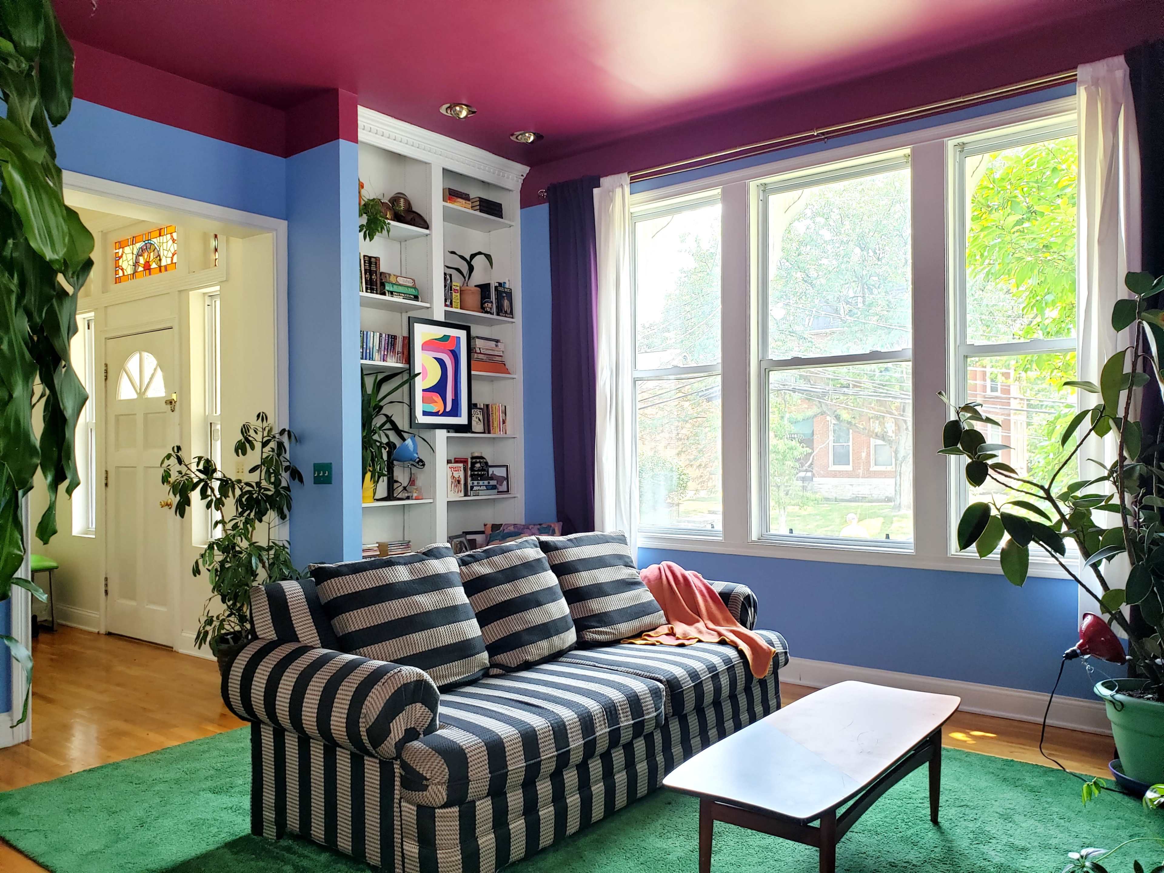 A striped sofa with a colorful throw rests on a green carpet in a brightly lit living room featuring large windows and houseplants.