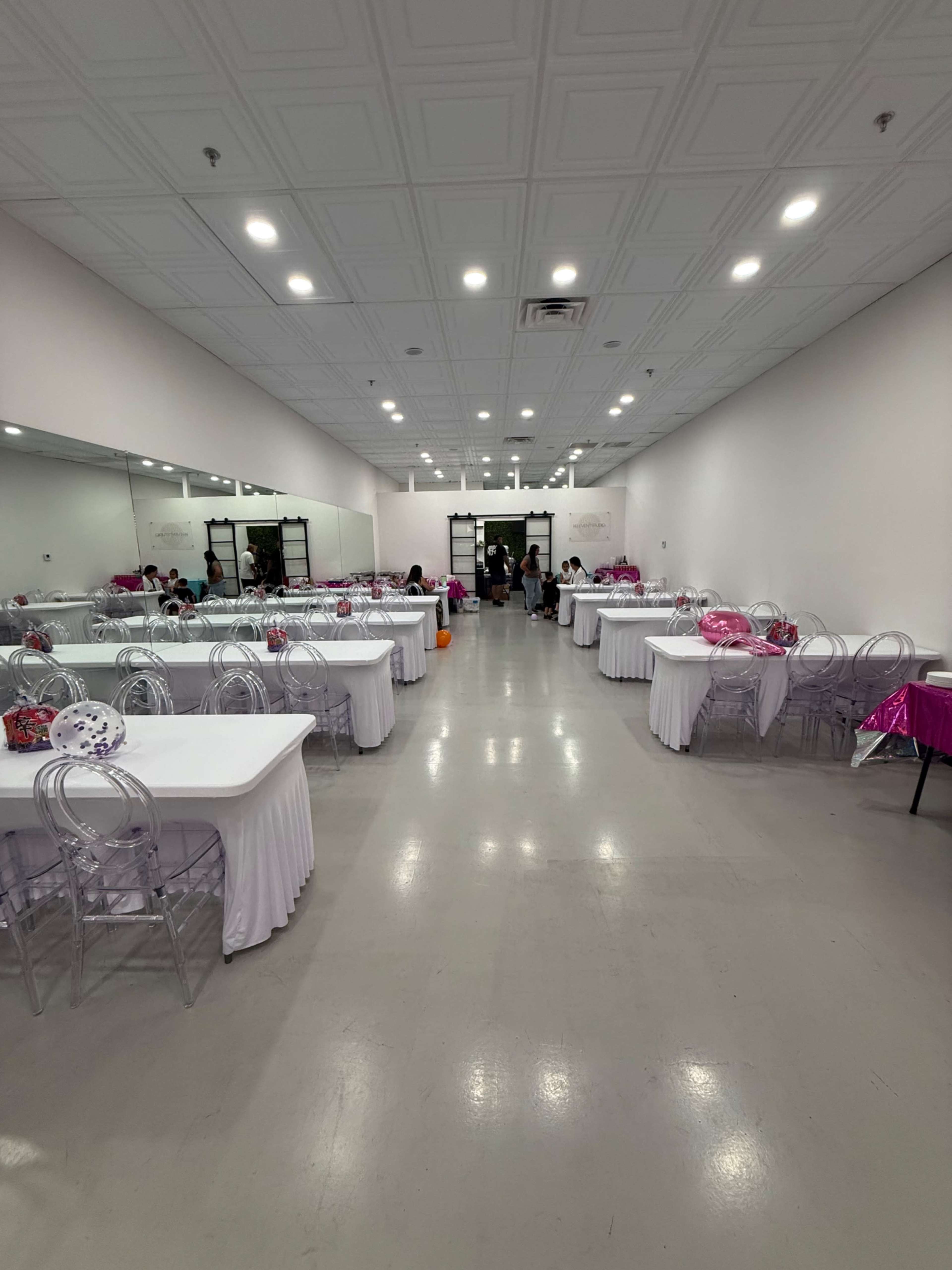 The image shows a spacious room set up for an event, featuring multiple long tables covered with white tablecloths and transparent chairs, with guests mingling in the background.