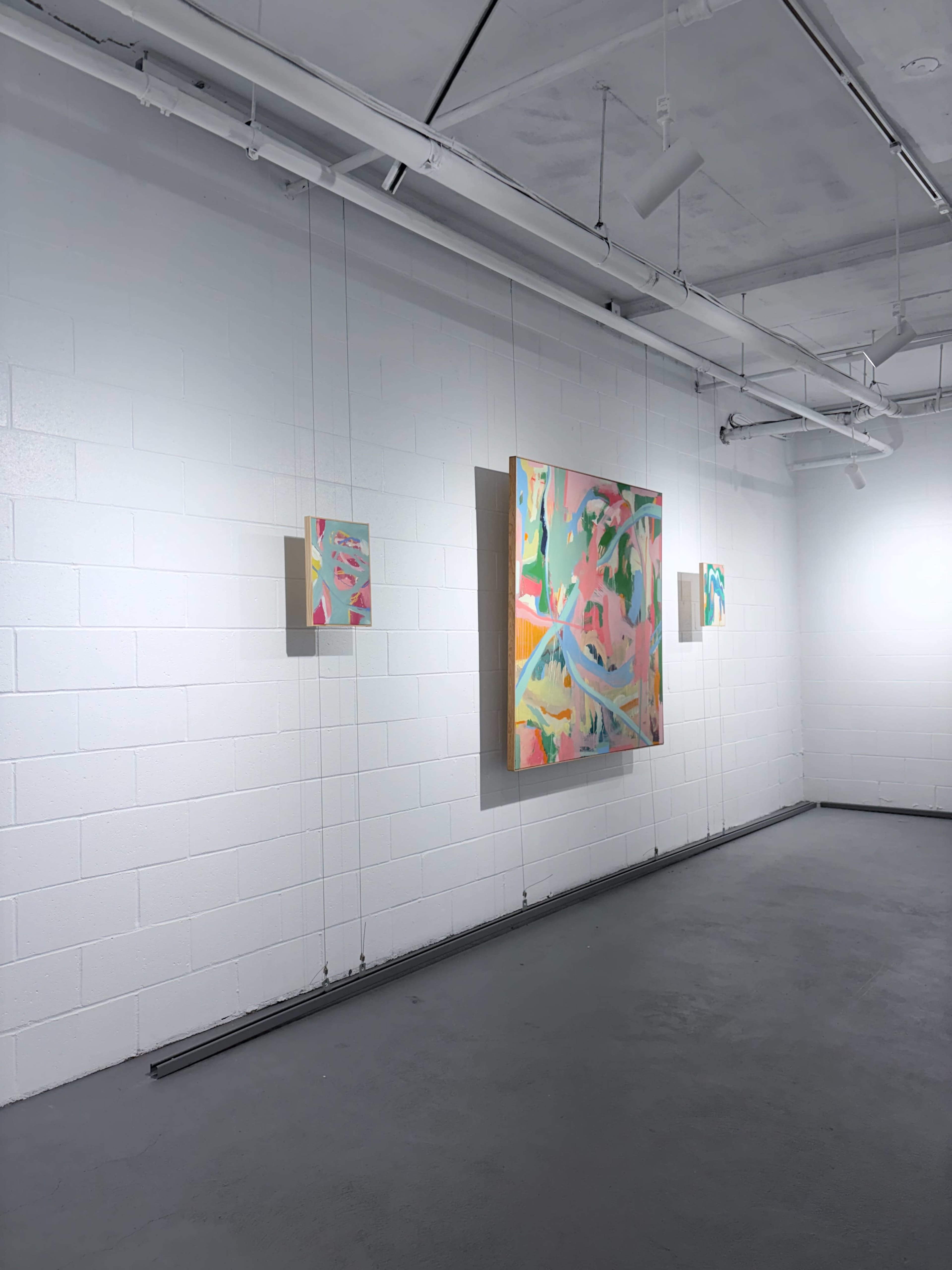 The image shows an art gallery interior with three colorful abstract paintings displayed on a white wall.
