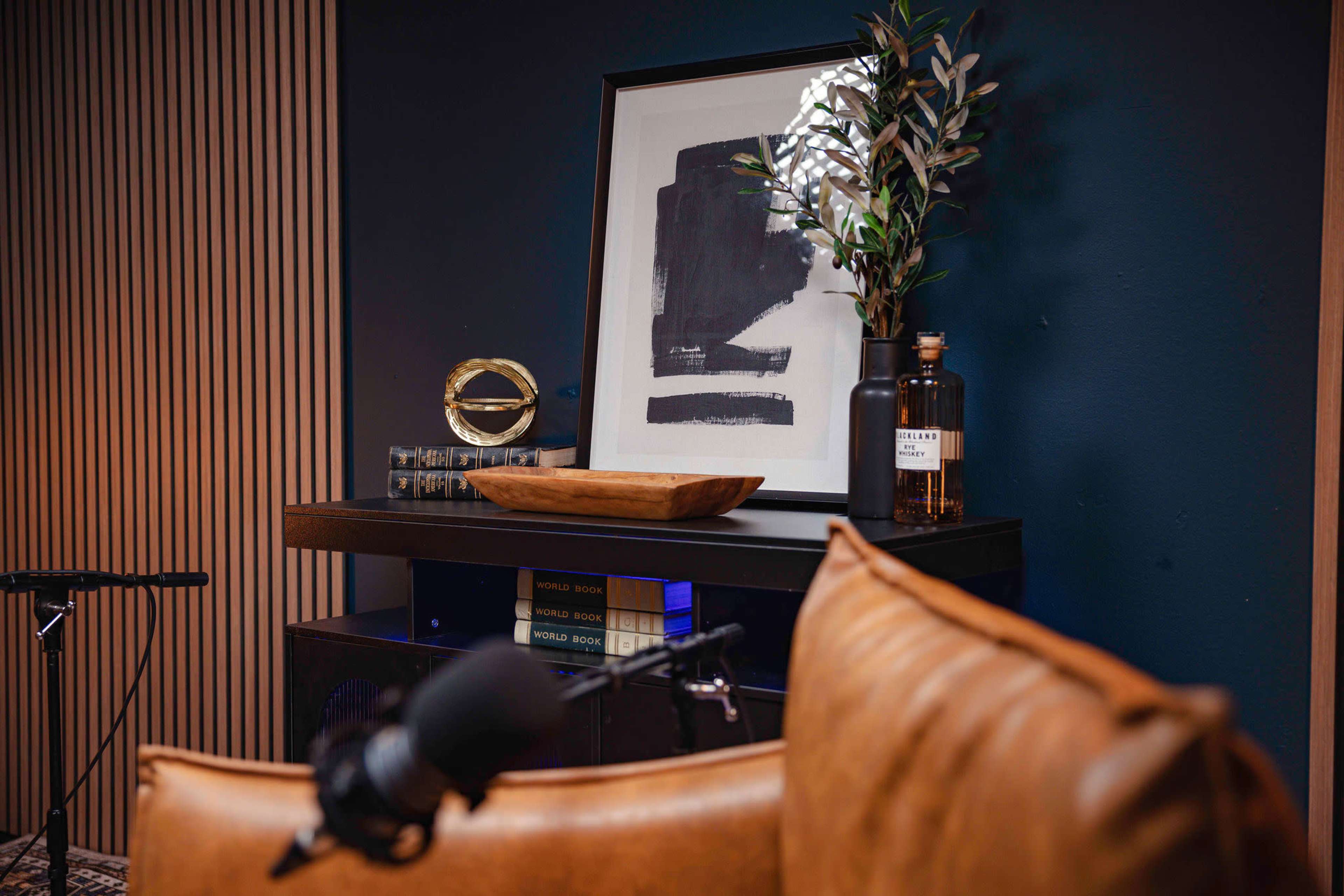 A brown leather couch is positioned in front of a dark blue wall, featuring a framed artwork, a decorative plant, and a bottle on a wooden shelf.