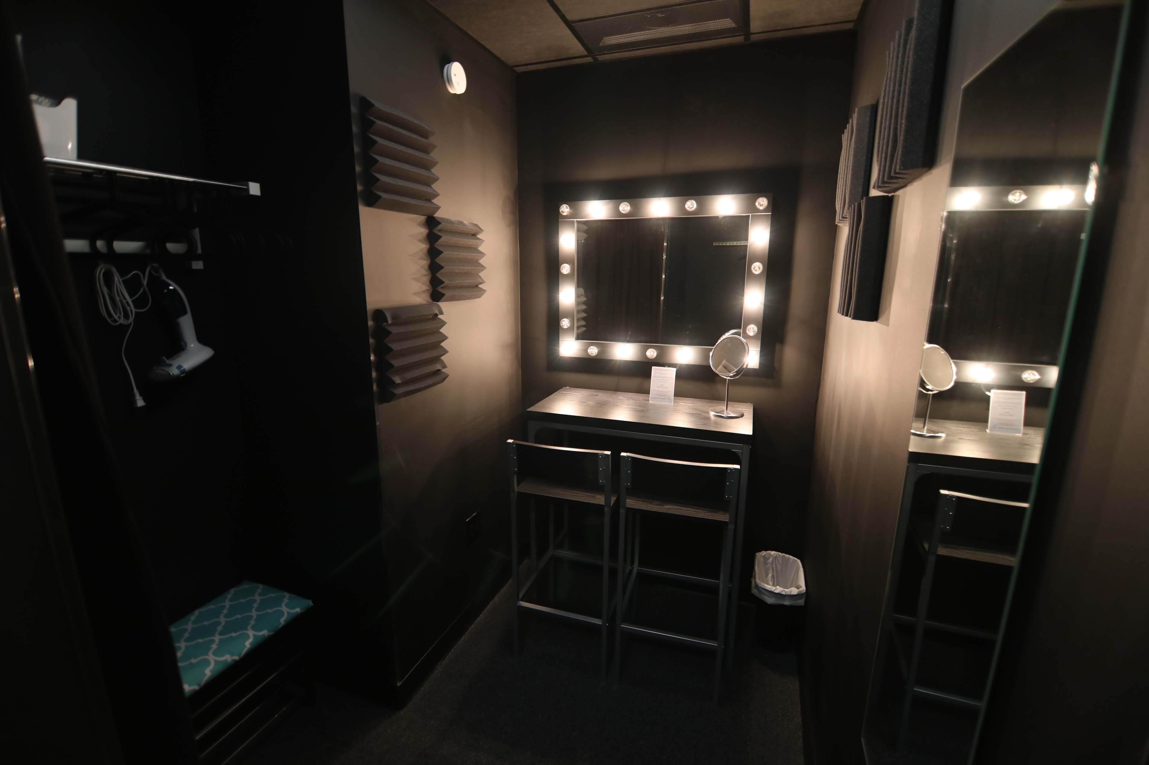 A small, dimly lit room with a mirror surrounded by bright bulbs, a table with two stools, and a closet space on the left.