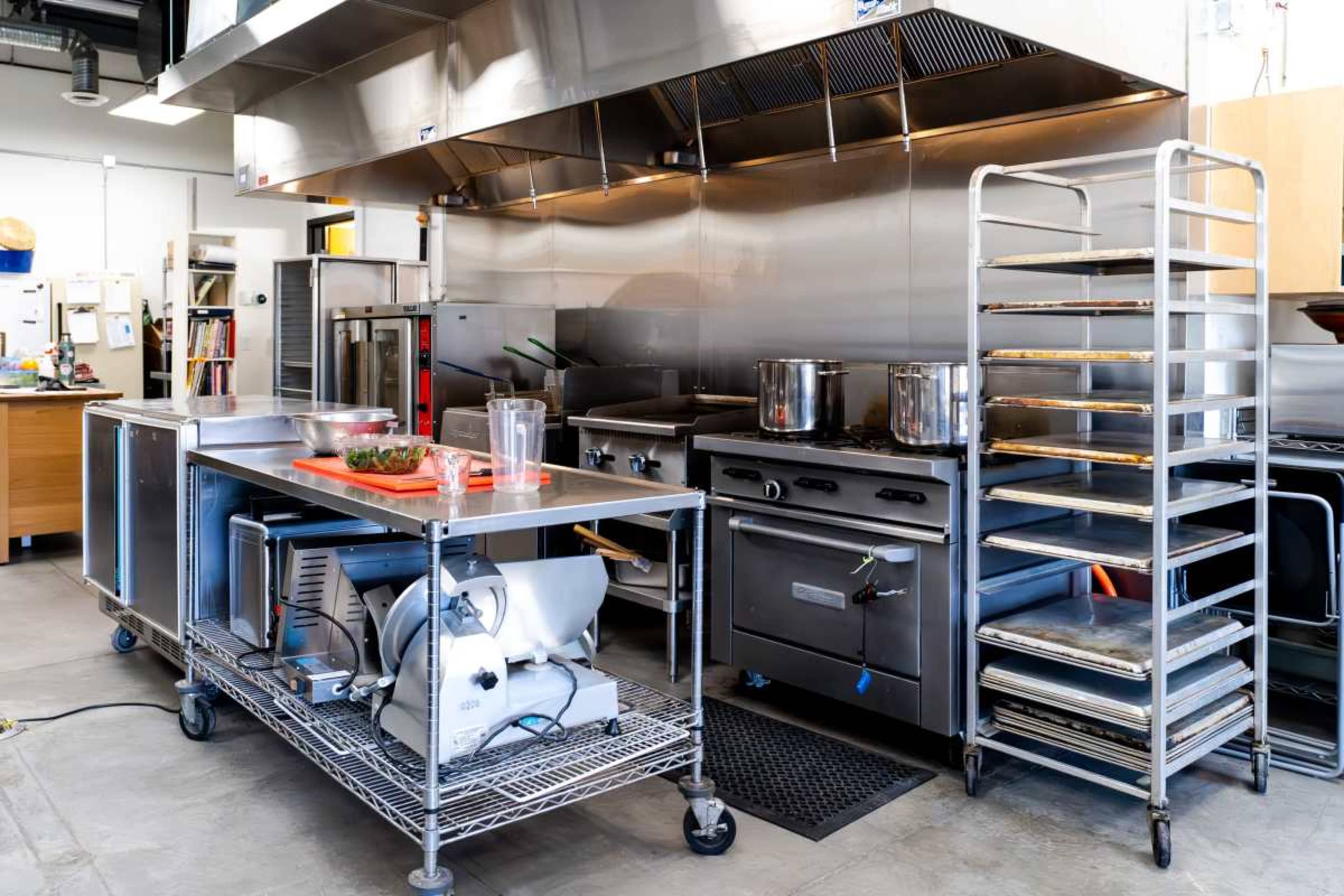 The image shows a commercial kitchen equipped with stainless steel appliances, cooking stations, and a rack for storing cookware.