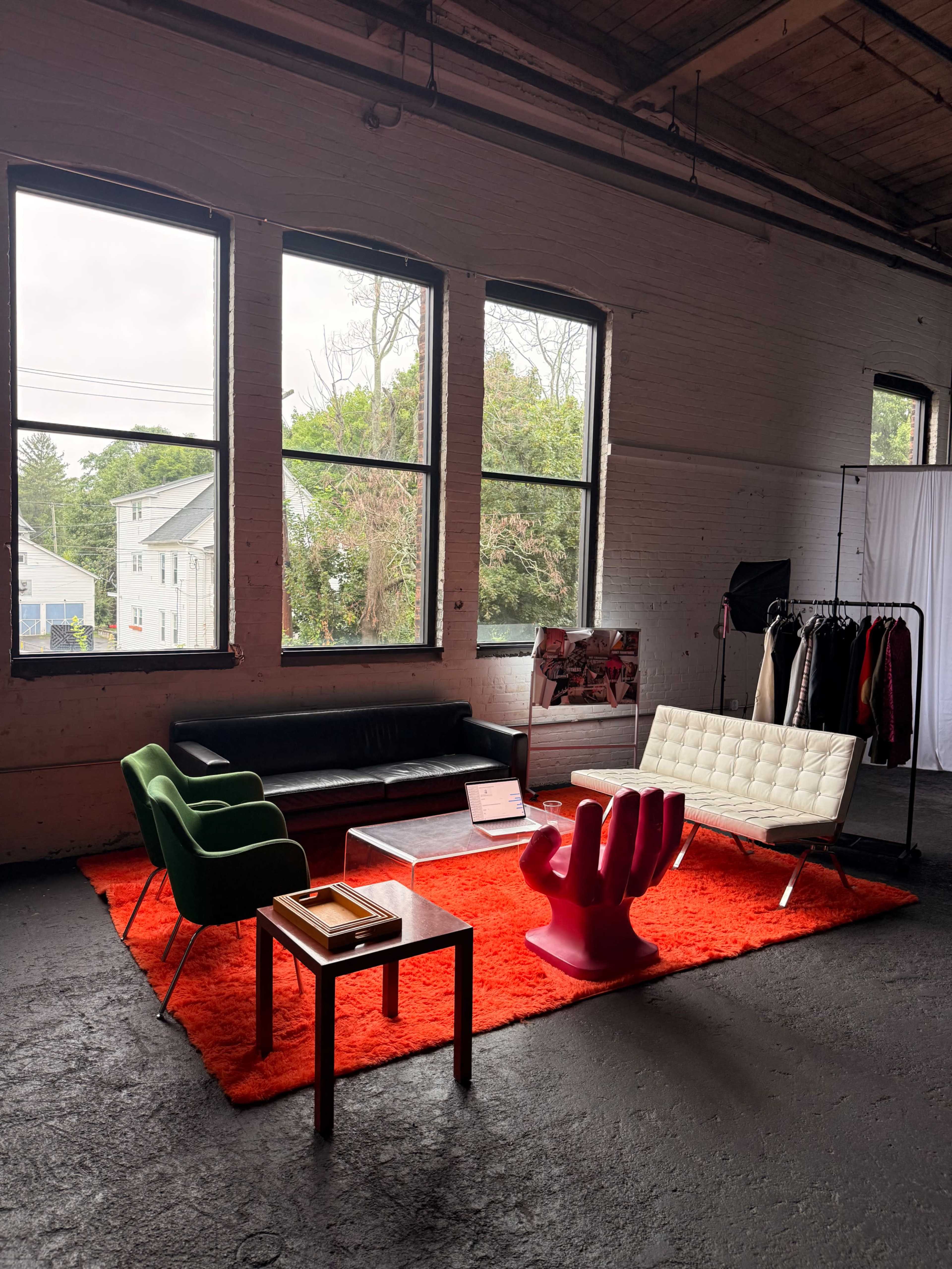 The image shows a minimalist room with large windows, featuring a black sofa, a white chair, green and pink accent chairs, a glass coffee table, and a red rug.