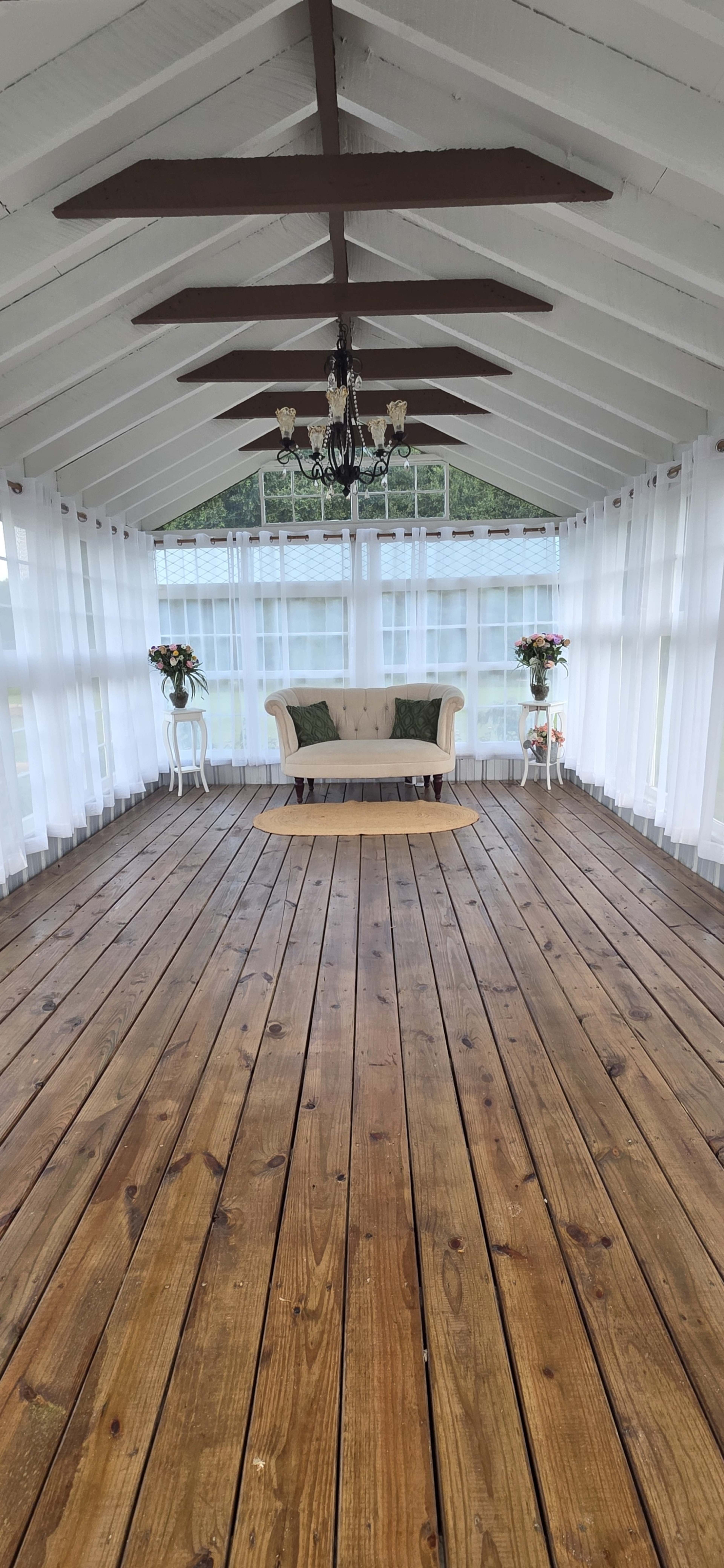 The image shows a spacious, well-lit room with wooden flooring, large windows, a central wooden platform, and a sofa against the wall, adorned with decorative plants on both sides.