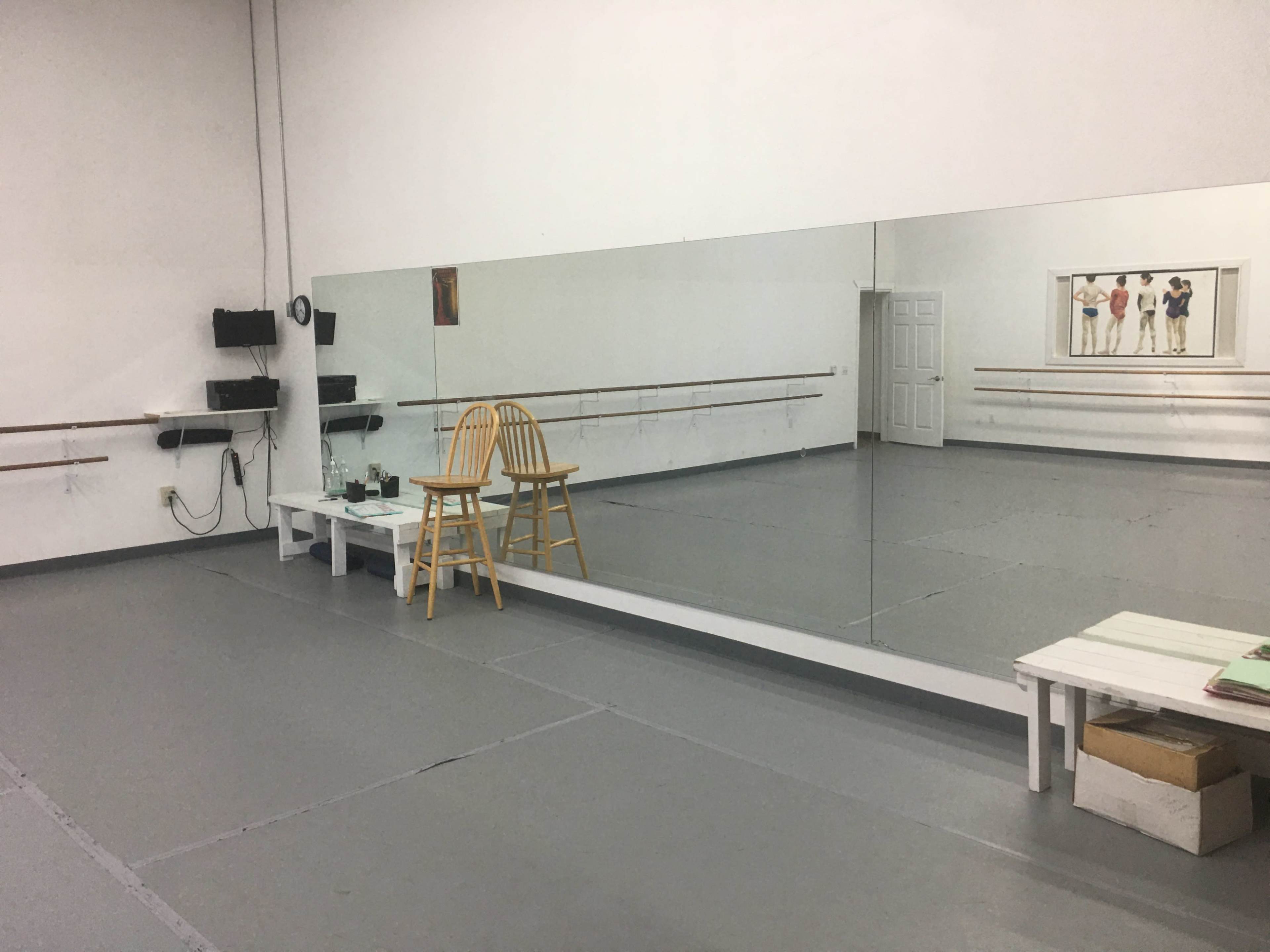 An empty dance studio features a large mirror, wooden barre, a chair, and stacked boxes against the walls.