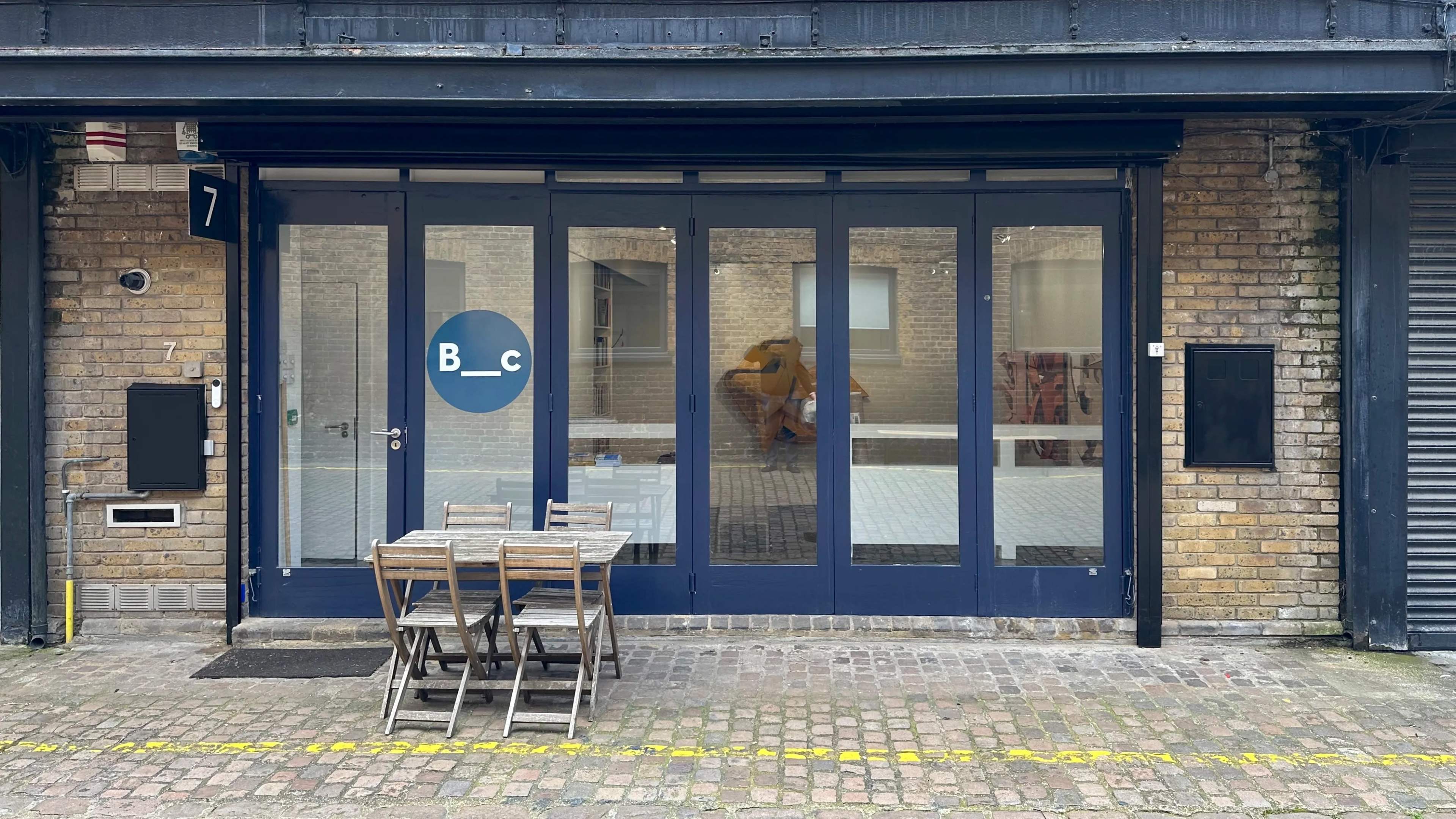 The image shows a modern storefront with large glass windows and a blue logo, accompanied by a small outdoor seating area with wooden chairs and a table.