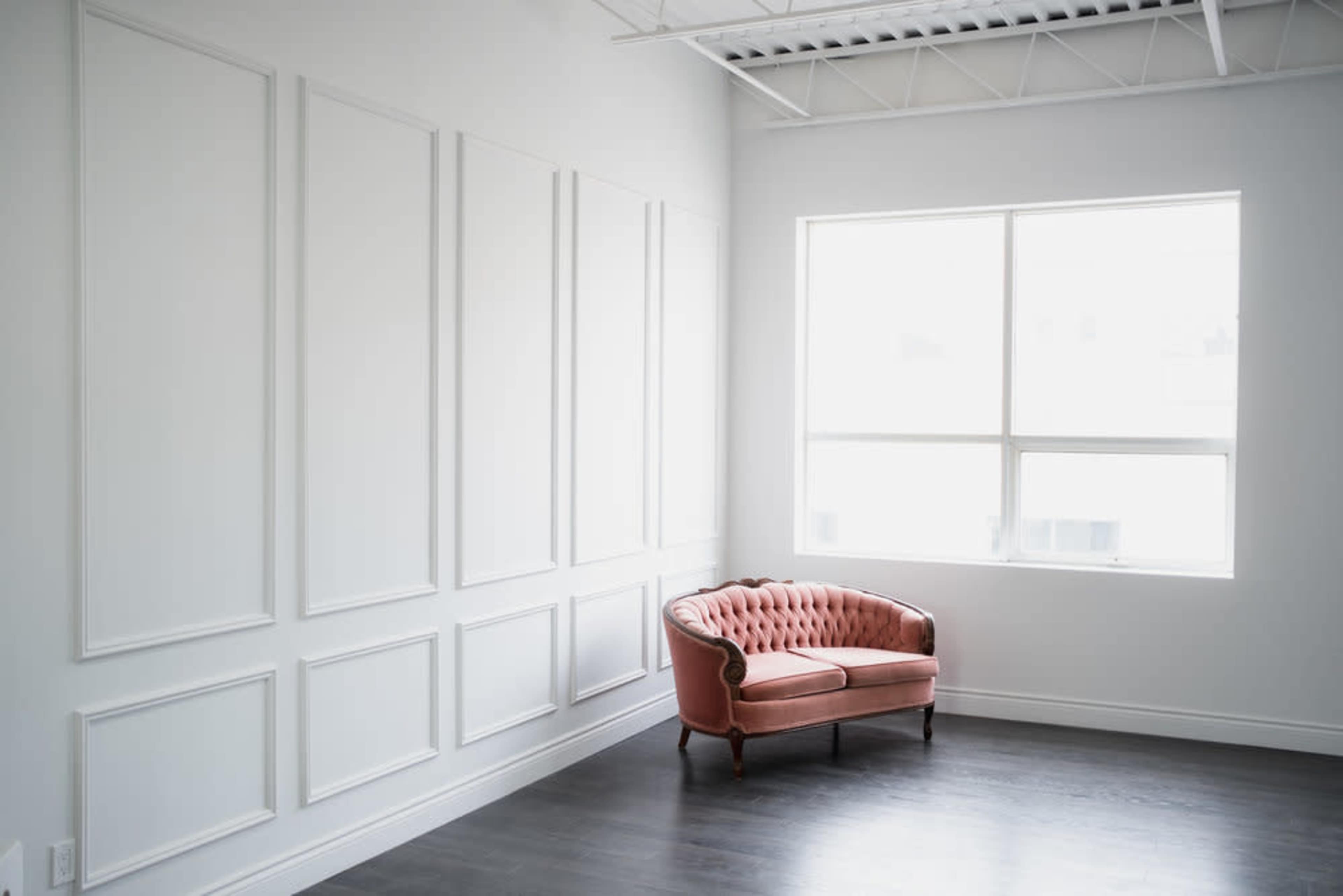 A pink tufted sofa is placed against a white paneled wall in a well-lit room with large windows.