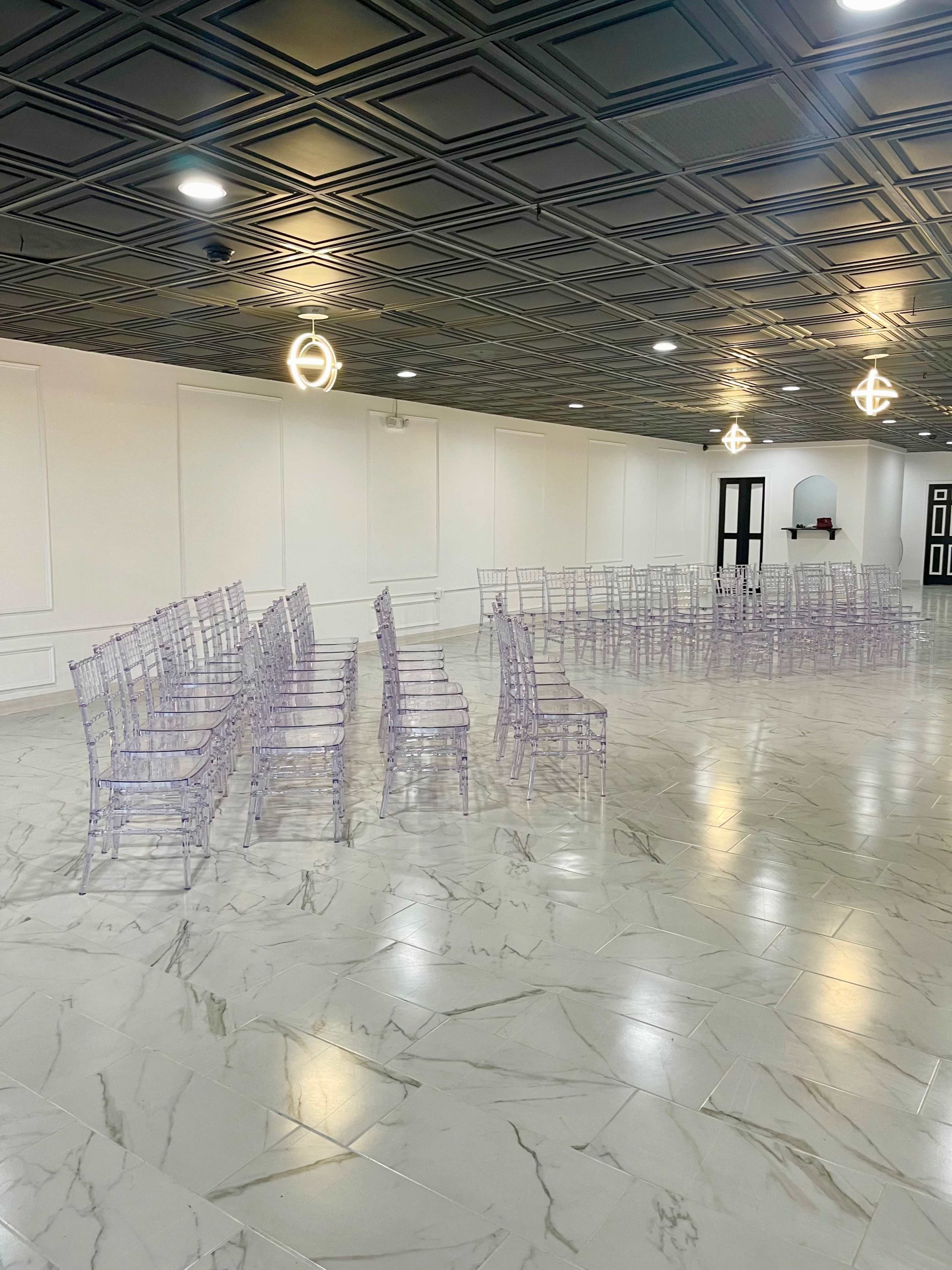 A spacious room features arranged transparent chairs on a marble floor beneath a patterned ceiling.