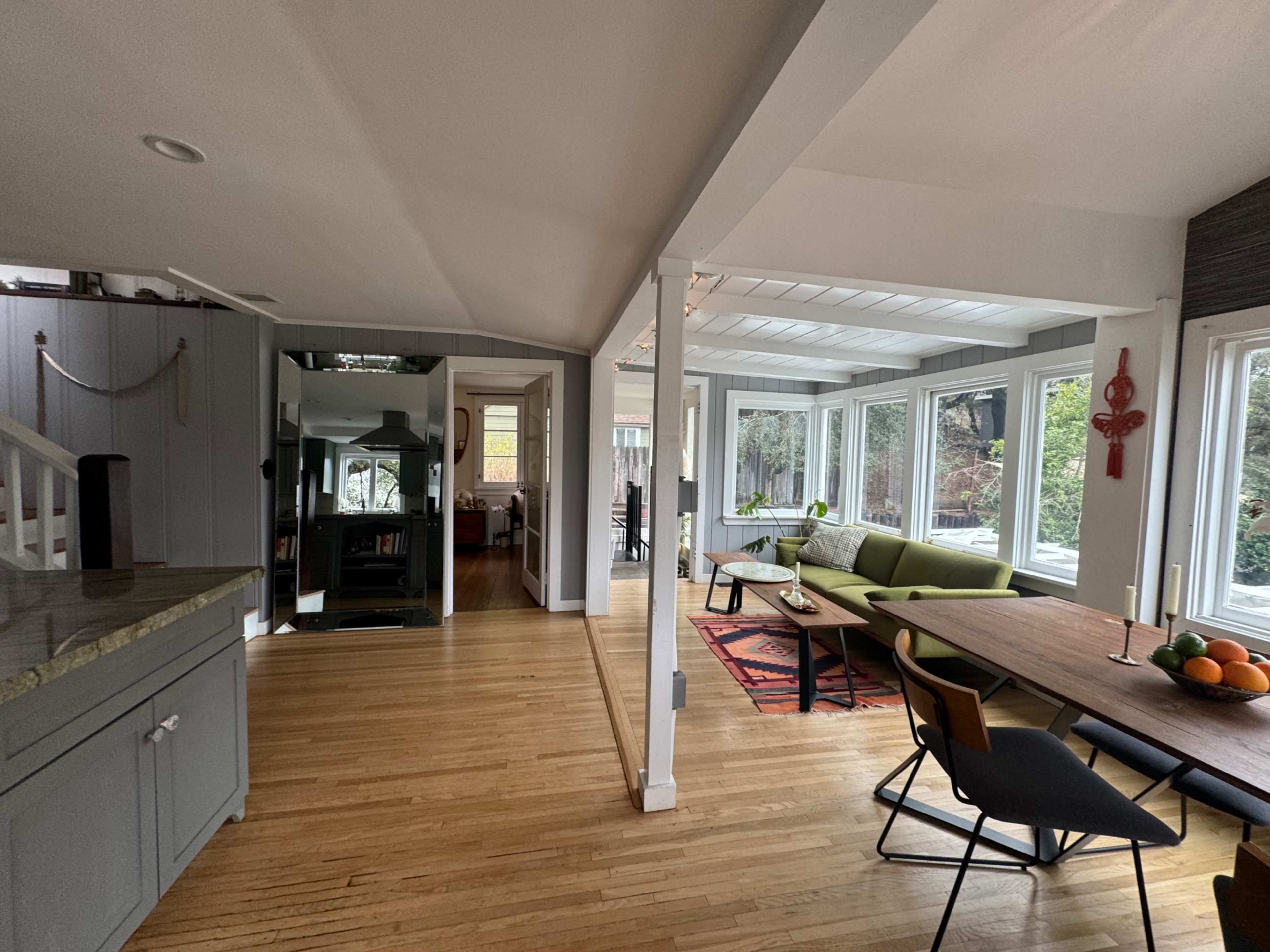 The image shows a modern interior space with an open layout, featuring a kitchen area, a dining table, and a sunlit sitting area.