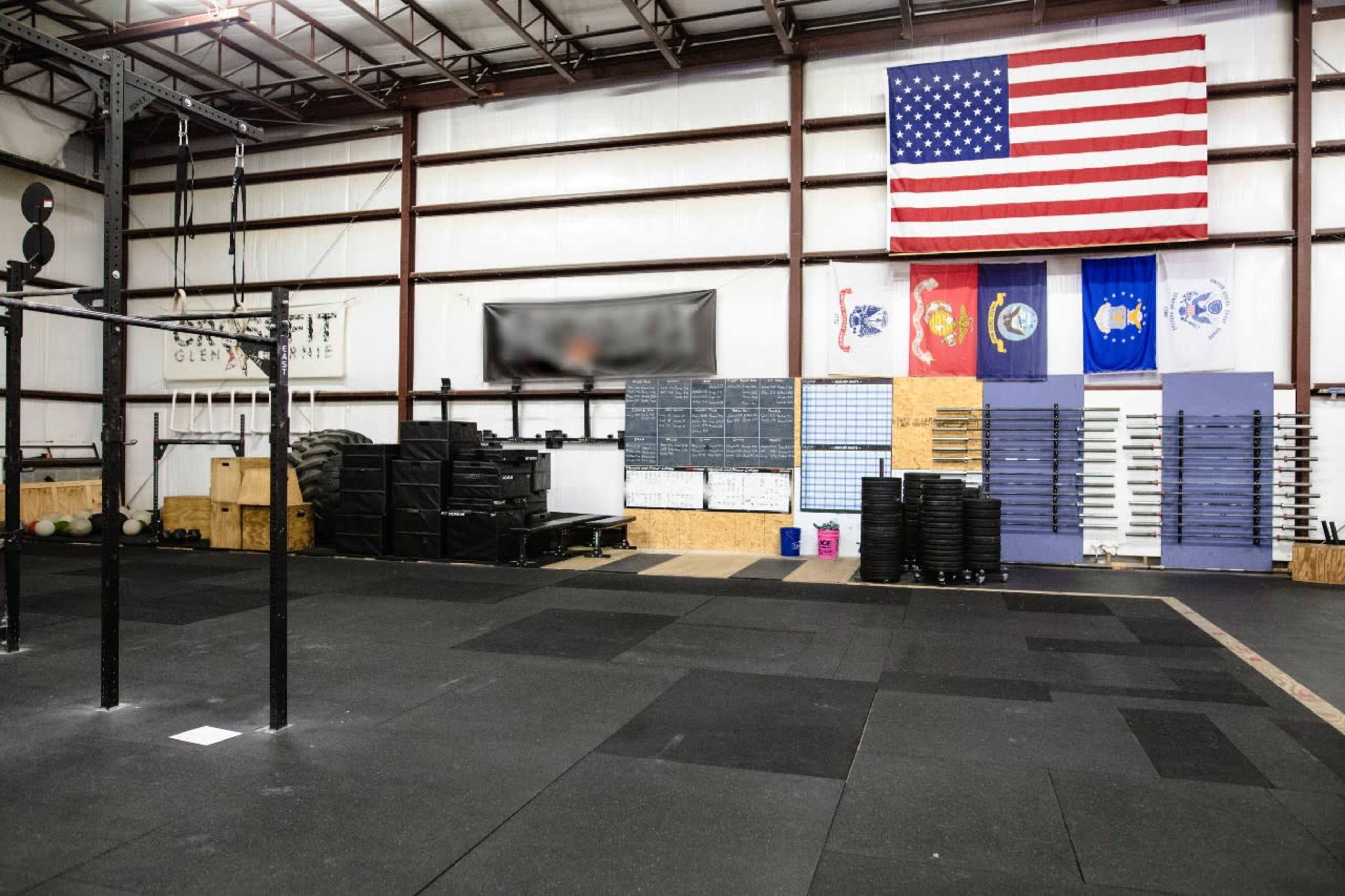 The image shows the interior of a gym featuring workout equipment, a rubber floor, and various flags hanging on the wall.