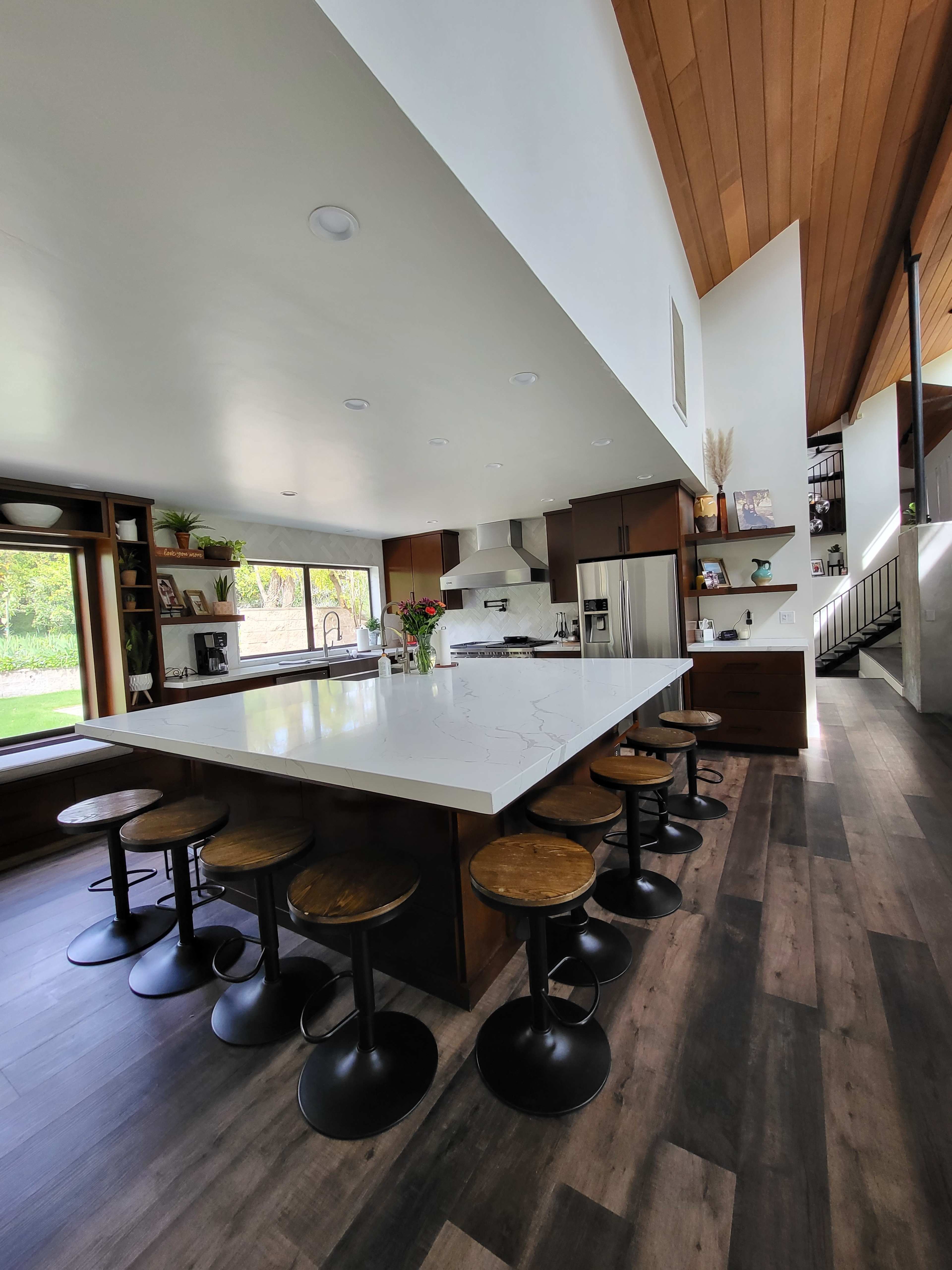 A modern kitchen with a large white island, dark cabinetry, and wooden bar stools arranged around it.