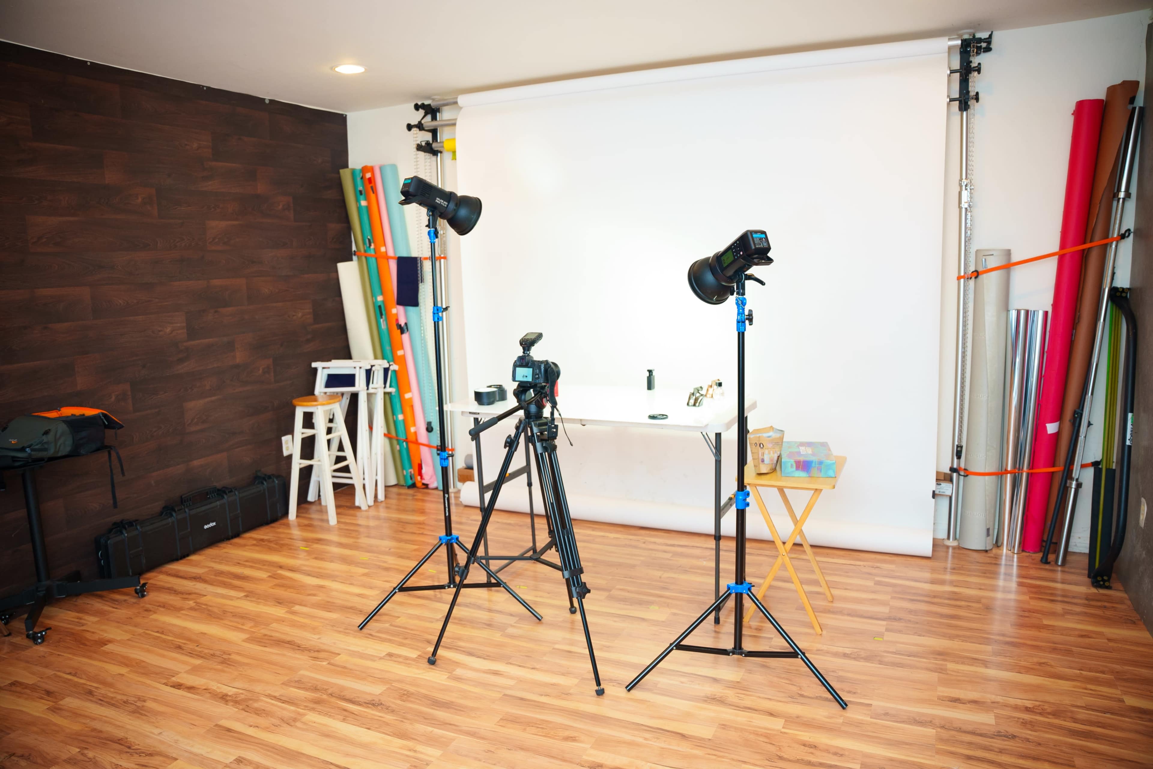 A photography studio is set up with two light stands, a camera on a tripod, and a table in front of a blank backdrop, surrounded by colorful photography materials.