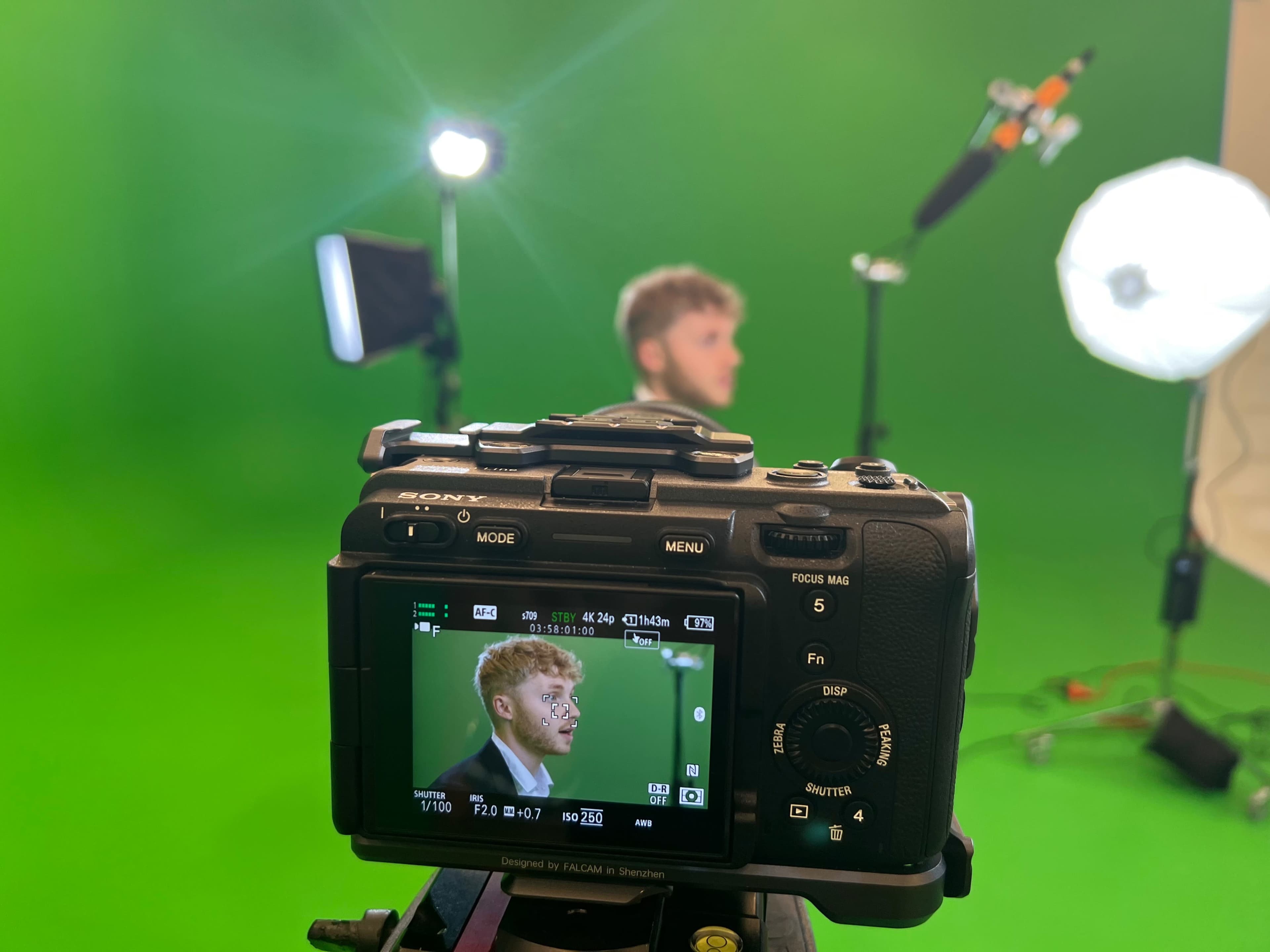 A camera is positioned on a tripod with its screen displaying a man in front of a green screen, while studio lights illuminate the scene.