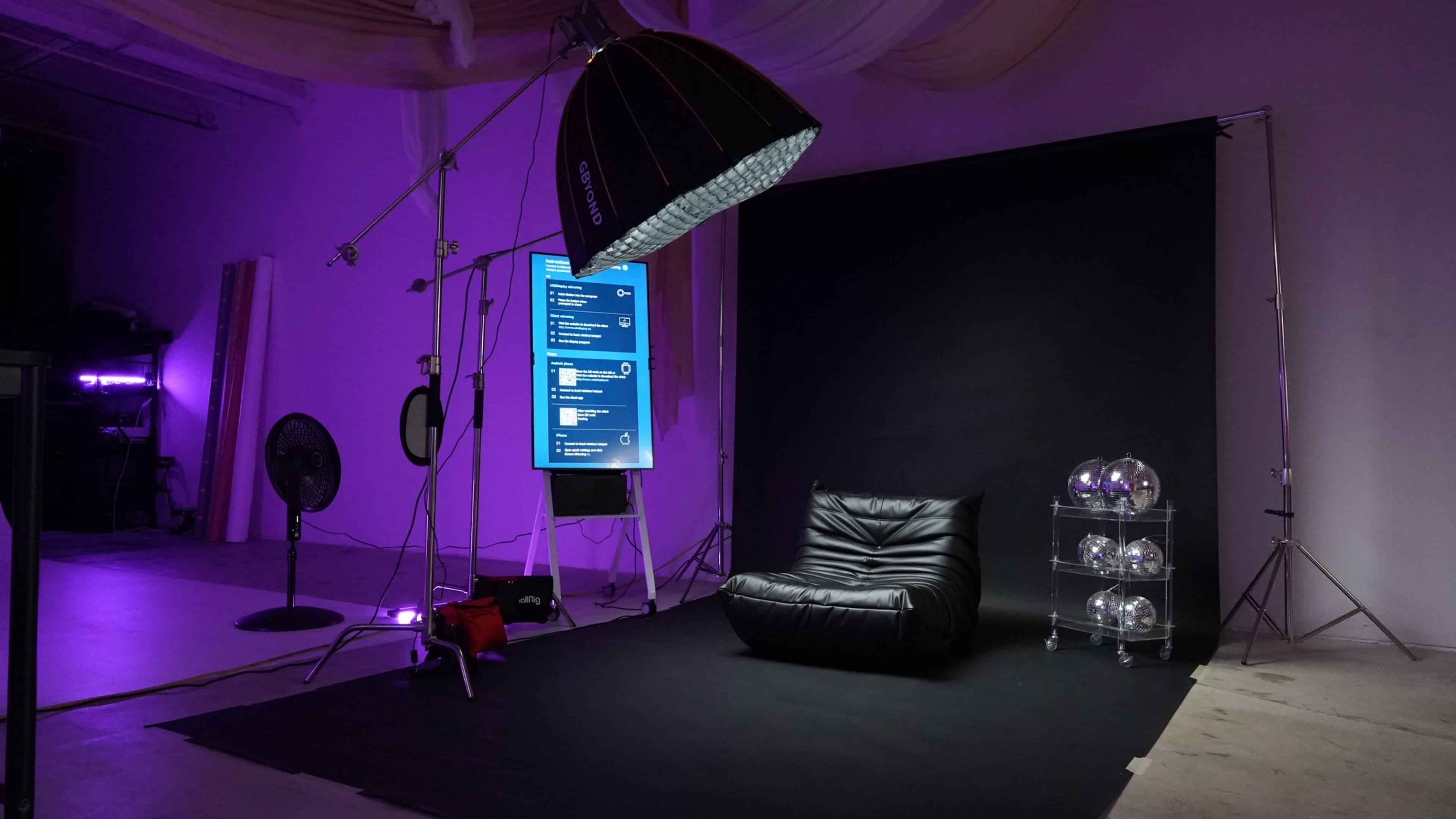 A photography studio setup with a black backdrop, a large softbox, a monitor displaying digital content, a black lounge chair, and a shelf with glass spheres.