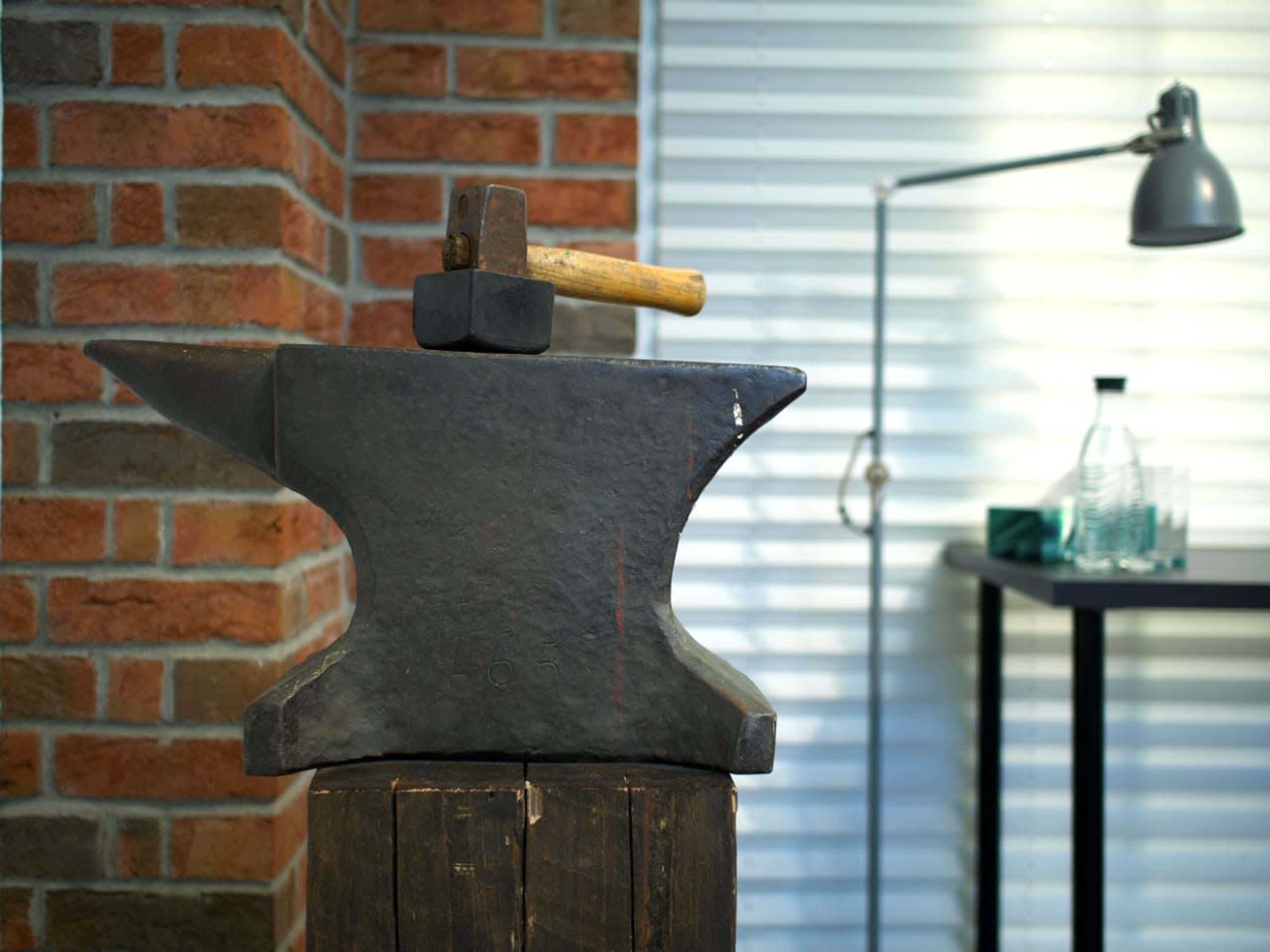 The image shows a blacksmith's anvil resting on a wooden stand, with a hammer placed on top, against a backdrop of brick walls and a nearby lamp.