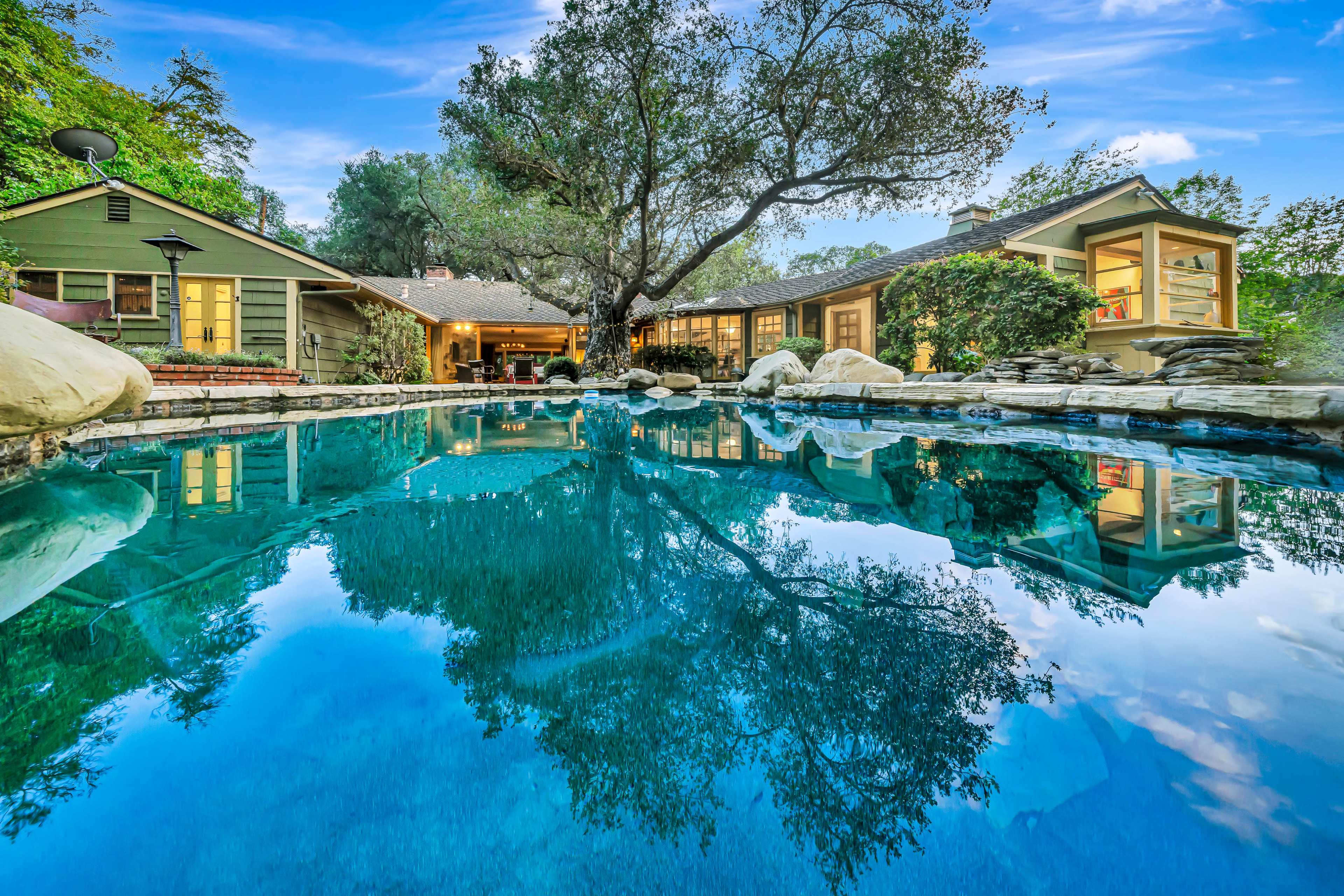 A tranquil outdoor scene features a swimming pool reflecting trees and a nearby house with large windows surrounded by lush greenery.